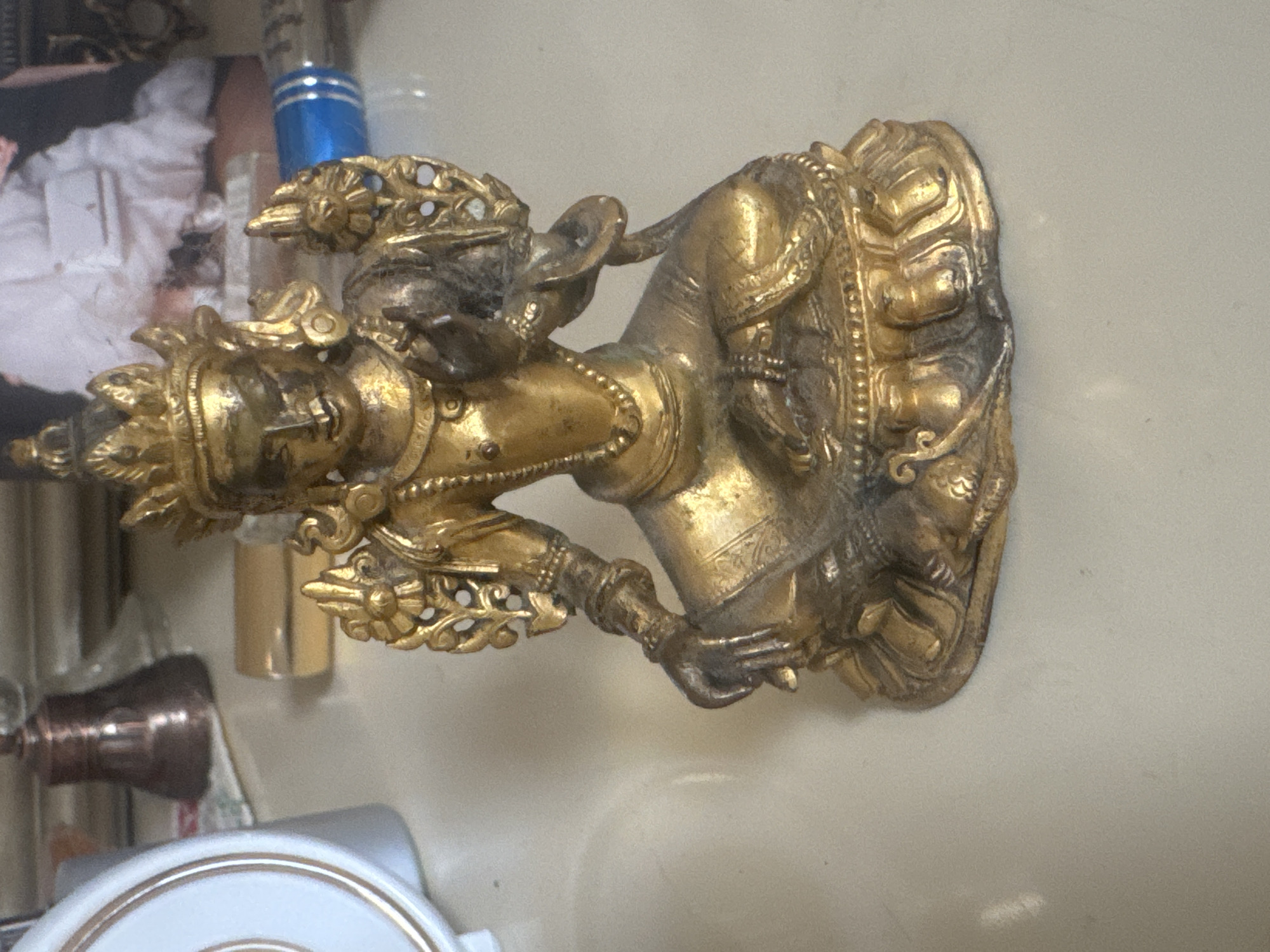 A gilded Buddha statue sits on a light-colored surface, possibly a table. The statue depicts a seated figure with a crown, elaborate jewelry, and intricate carvings. The background is blurred, showing a white ruffled garment, a blue object, and other indistinct items. The lighting suggests an indoor setting. There is no indication of the location being Sochi, Russia.