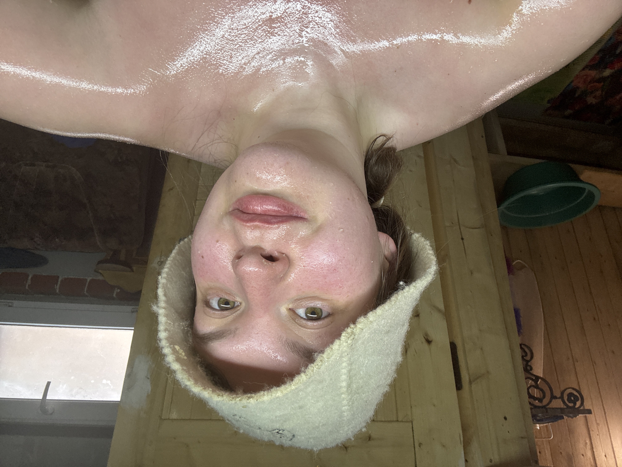 A sweat-covered person wearing a felt sauna hat is upside down in a sauna. The individual's face is flushed, and their skin is glistening with moisture. The sauna has wooden walls and a window. A green bucket and decorative metal item are visible in the background. There is no information on the city of Lukintsy, Russia.