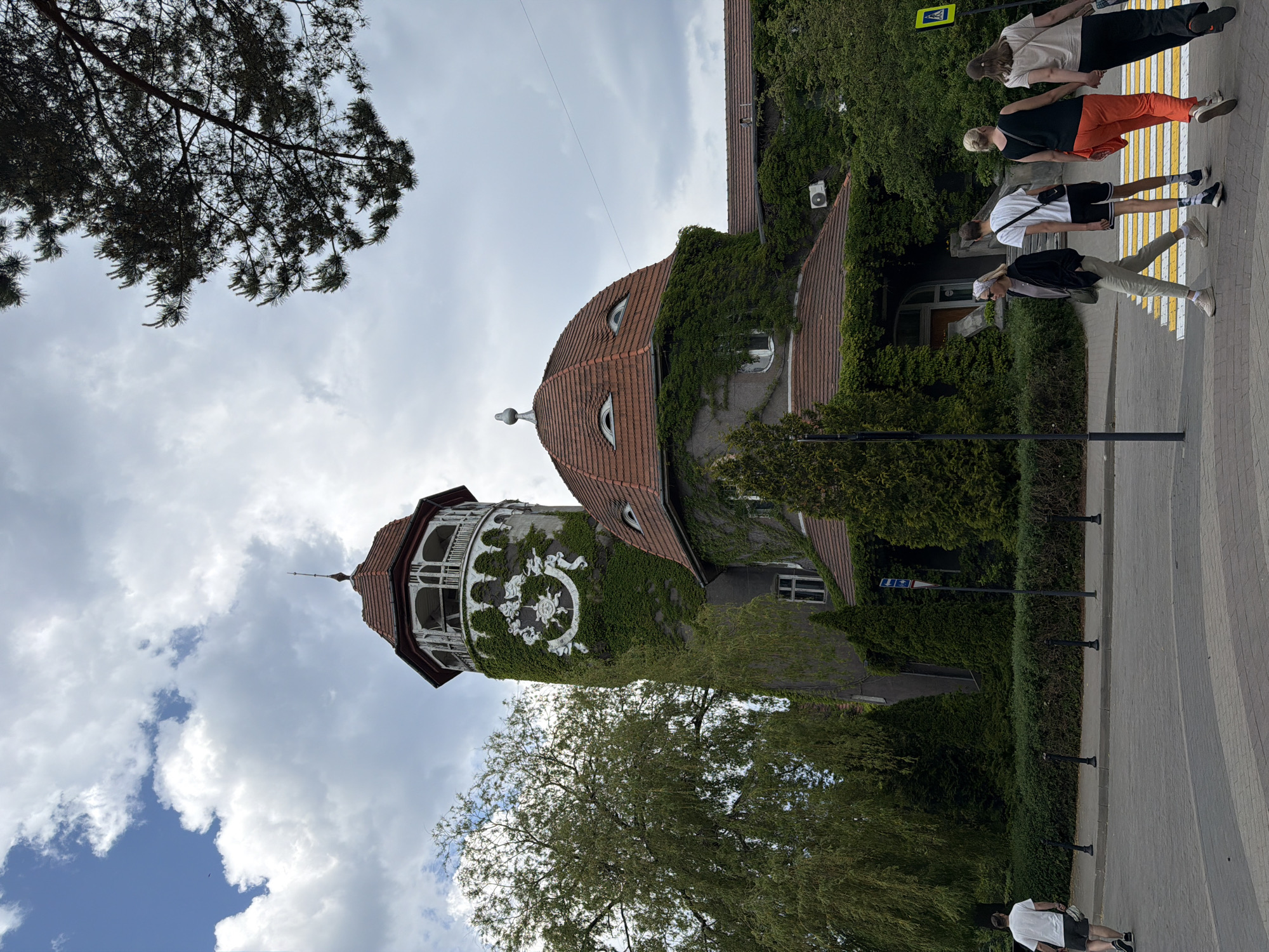 The image depicts the Svetlogorsk Water Tower and an adjacent building, commonly known as the "Magic Eye" building, in Svetlogorsk, Kaliningrad Oblast, Russia, under an overcast sky with visible patches of blue.
In the center-left, a tall, cylindrical stone tower, heavily covered in green ivy, rises with a reddish-brown conical tiled roof topped by a small finial. Below the roofline, a white, open-air octagonal balcony with decorative railings encircles the tower. Beneath the balcony, a large, ornate white crest-like decoration is prominently displayed against the ivy-covered stone. To the right of the tower, a lower, domed building with a matching reddish-brown tiled roof is also covered in ivy. This dome features two semi-circular dormer windows that resemble eyes, and a small finial ornament at its apex. Further right, another section of the building with a red-tiled roof extends, also draped in ivy, with an external air conditioning unit mounted on its wall.
A large, light-green deciduous tree stands to the left of the tower, and various evergreen shrubs and trees line the foreground and surround the buildings. The street in the foreground features a yellow and white striped pedestrian crosswalk. Black bollards separate the pavement from the roadway. A dark lamppost stands near the crosswalk. A blue traffic sign is visible behind the bollards on the right, and a yellow pedestrian crossing sign is present on the far right.
Several individuals are present in the foreground:
On the far left, a male in a white t-shirt and dark shorts walks away from the viewer on the pavement, carrying a white bag.
On the right, four individuals cross the pedestrian walkway from left to right. The leftmost of this group is a female in a black jacket, light trousers, and a light headscarf. Next to her, a male in a white t-shirt and dark shorts with white stripes walks away from the viewer. To his right, a female in a black sleeveless top and orange trousers walks away, holding hands with the female next to her, who is dressed in a light-colored top and dark trousers.
