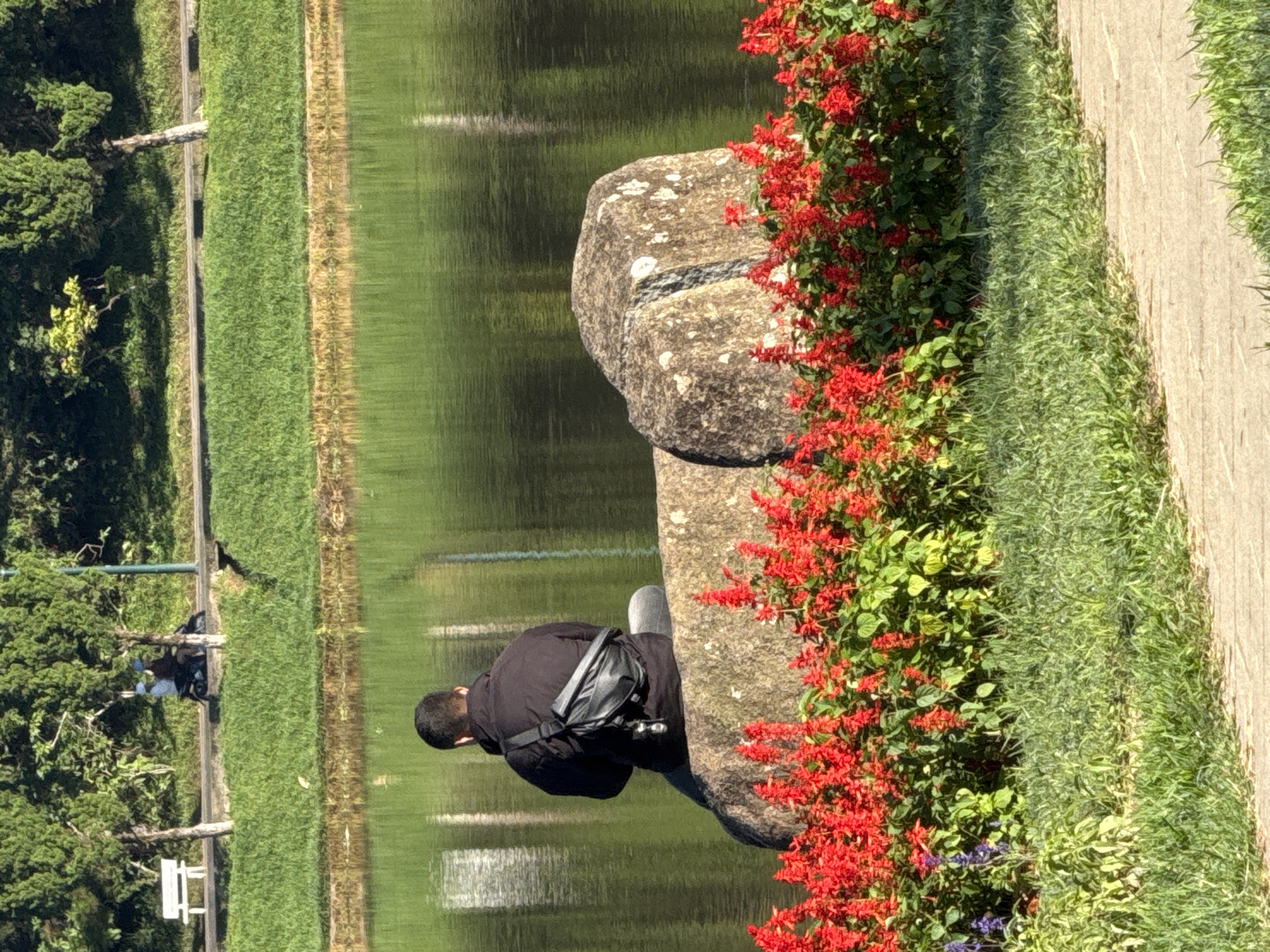 A man wearing a black jacket and a dark crossbody bag is sitting on a large, rough-textured rock next to a body of water. Red flowers are blooming in front of the rock, and green grass is visible in the foreground. In the background, there is a path with trees and two people riding a motorcycle. The scene appears to be in a park or by a lake in Da Lat, Vietnam, as indicated by the presence of a white bench, lush greenery, and the overall serene atmosphere characteristic of the city's landscapes.