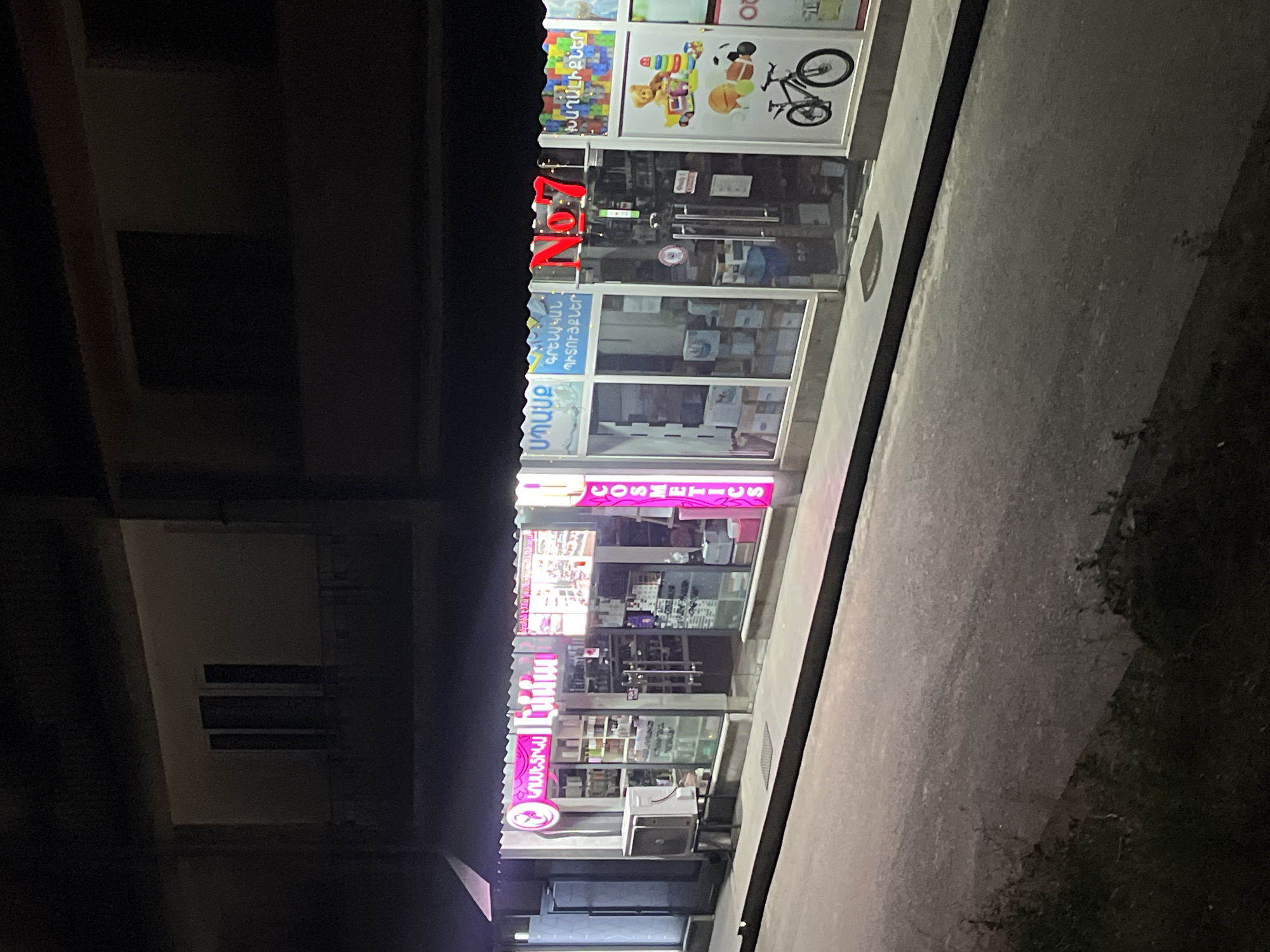 The image shows the storefront of a commercial building at night, likely in Gladzor, Armenia. The facade features multiple shops with illuminated signs. One prominent sign, in red neon letters, reads "ZON". To the left of this sign, another sign in Armenian script, with bright pink lettering, appears to advertise "COSMETICS". Below the "ZON" sign, more windows display the interiors of shops, revealing shelves stocked with various products. A white air conditioning unit is mounted on the exterior wall beneath the cosmetics sign. The building is set against a dark background, suggesting it is nighttime. The street or sidewalk in front of the building is visible and appears to be made of concrete or asphalt. The overall impression is of a well-lit commercial street at night.