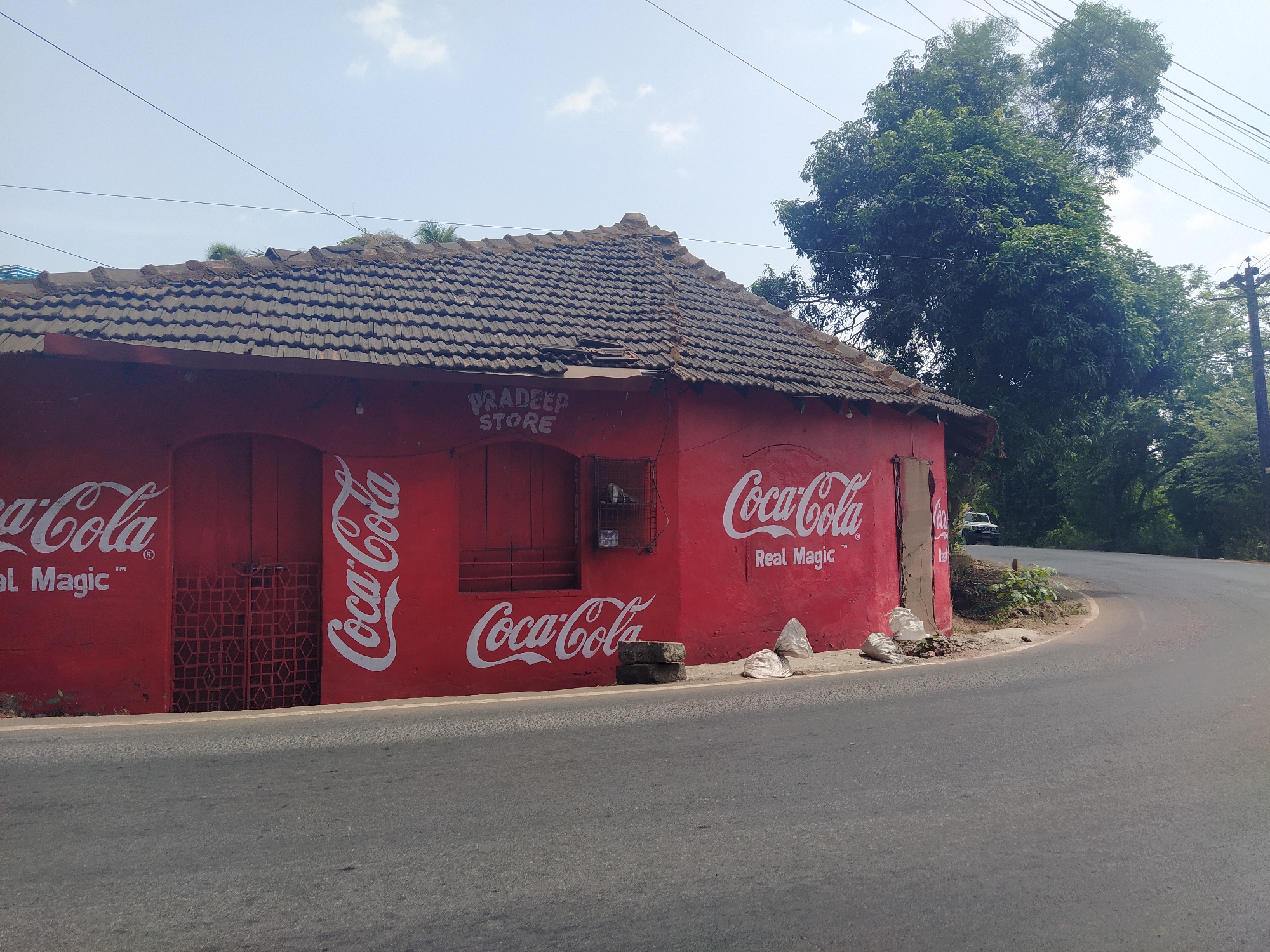 A red-painted building with a tiled roof stands adjacent to a road. The building prominently features "Coca-Cola" branding on its exterior walls along with the inscription "Real Magic." Above a window, the words "PRADEER STORE" are visible. The roof shows some disrepair. A road curves around the side of the building, and trees are visible in the background. The location is Moira, India. The scene appears to be during daylight hours.