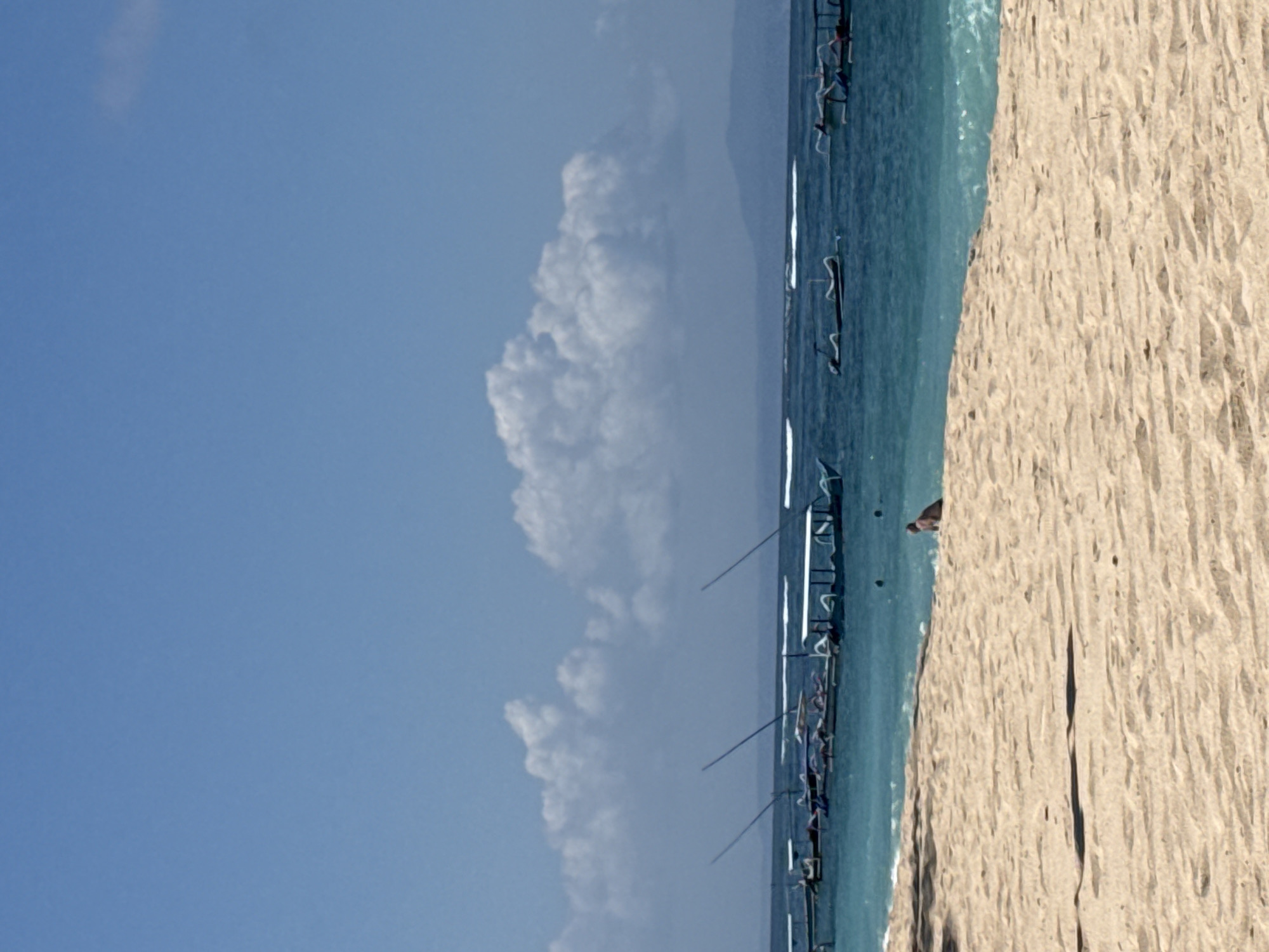 This image depicts a tropical beach scene, likely in Kuta Selatan, Indonesia, with a focus on the coastline and the sea. The foreground is dominated by a sandy beach with subtle undulations and shadows suggesting a bright, sunny day. In the middle ground, the turquoise waters of the ocean are visible, with gentle waves breaking near the shore. A line of traditional outrigger boats with canopies are moored or sailing in the middle distance, some with their masts raised. In the background, the ocean extends to the horizon, meeting a hazy, mountainous landscape under a clear blue sky punctuated by scattered white clouds. A single person is visible in the water, appearing to be swimming or wading.