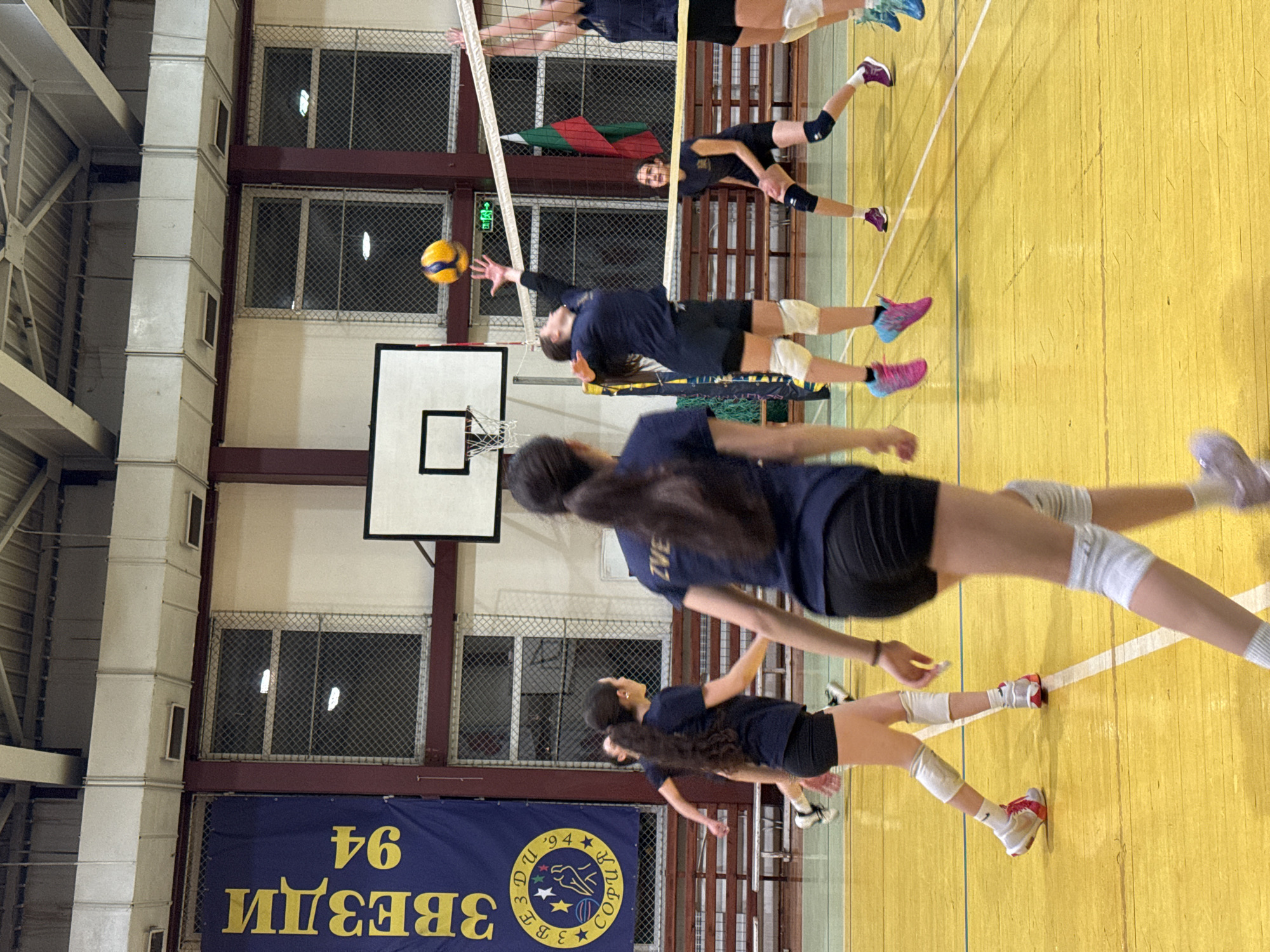 The image shows an indoor volleyball court in Sofia, Bulgaria, likely during a practice session. Several female volleyball players are visible, wearing matching navy blue shirts and dark shorts. One player is in mid-air, reaching out to hit a yellow volleyball over the net. Other players are positioned on the court, presumably preparing for their next move. The court has a yellow floor with white lines. A basketball hoop is visible on a nearby wall. A banner with the words "ЗВЕЗДИ 94" (Zvezdi 94) and the city name "СОФИЯ" (Sofia) is displayed, indicating the team and location. A Bulgarian flag is visible near the net. The background includes windows with wire mesh and support structures.