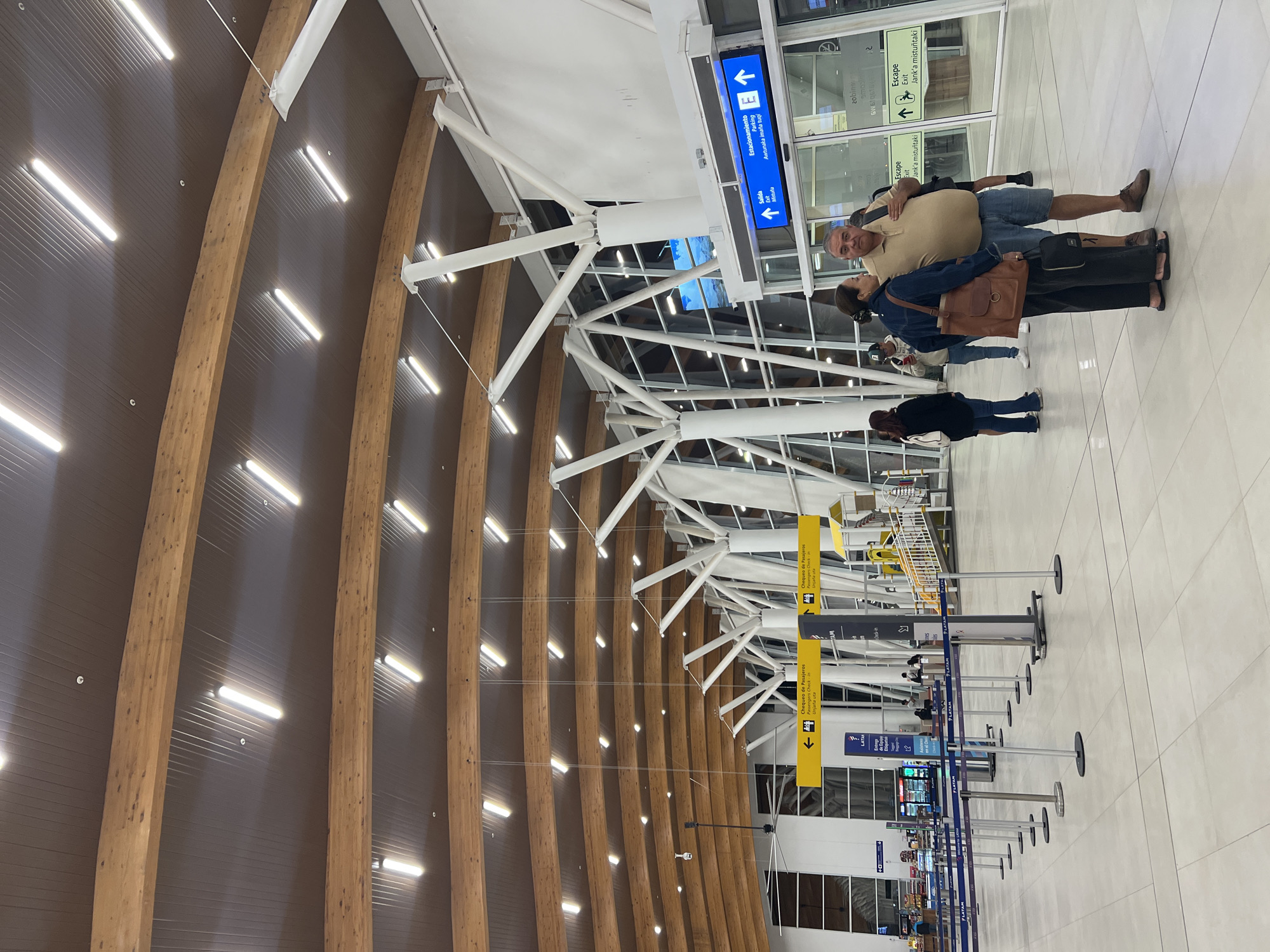 The image shows the interior of an airport terminal, potentially the Chacalluta International Airport in Arica, Chile, based on the signage. The terminal features a distinctive ceiling design with curved wooden beams and integrated lighting. White structural supports connect the ceiling to the building's frame.
In the foreground, there are queue barriers and signage directing passengers to "Chequeo de Pasajeros" (passenger check-in) and "Salida" (exit). The signs are in Spanish and Aymara, reflecting the local indigenous language.
A couple is standing near an exit door; the man is wearing a beige shirt and blue denim shorts, while the woman is wearing a blue jacket and black pants. Other individuals are also visible in the background, moving through the terminal.
The environment appears clean and well-lit, indicative of a modern airport facility.