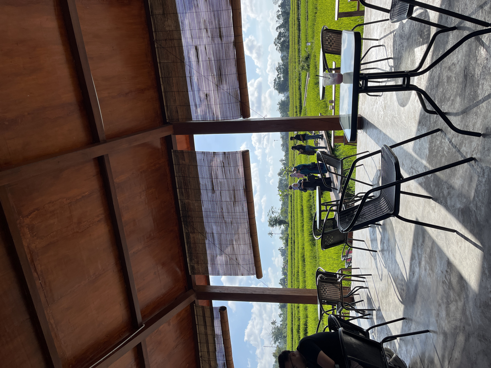 The image captures an outdoor cafe setting overlooking a vibrant green rice paddy field. The cafe structure features a wooden roof supported by pillars, with bamboo blinds covering parts of the openings. Several black metal tables and chairs are arranged on a concrete floor, some casting long shadows due to the sunlight. In the foreground, a person wearing a black t-shirt is seated at a table. In the middle ground, a group of people, some wearing headscarves, are walking on a wooden pathway within the rice field. The background reveals the expansive green rice terraces under a bright blue sky with scattered white clouds. The location is identified as Oro-oro Ombo, Indonesia, suggesting a rural or semi-rural environment known for its agriculture.