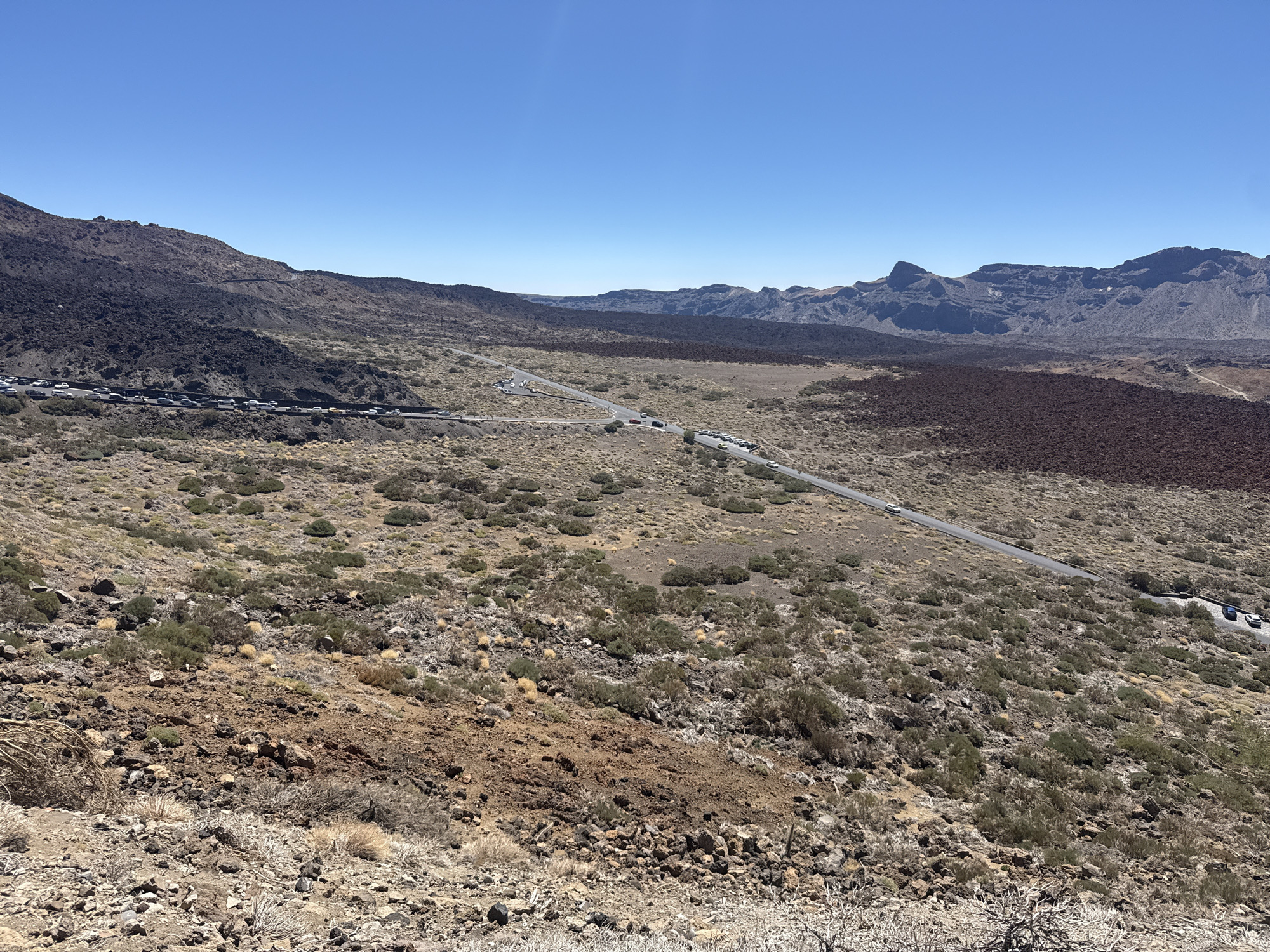 The image captures a panoramic view of a volcanic landscape under a clear blue sky. The terrain is rugged and arid, characterized by dark volcanic rock formations and sparse, low-lying vegetation. A winding road cuts through the landscape, with several cars visible on it. The road appears to be a popular route, as evidenced by the lines of parked cars along its edges in some sections, suggesting it might be a scenic viewpoint or a point of access to hiking trails. In the distance, majestic mountains form the horizon, their contours softened by atmospheric perspective. The overall impression is one of a dramatic and remote natural environment, typical of volcanic national parks. This scene is located in the Teide National Park, which is situated on the island of Tenerife, in the municipality of La Orotava, Spain.