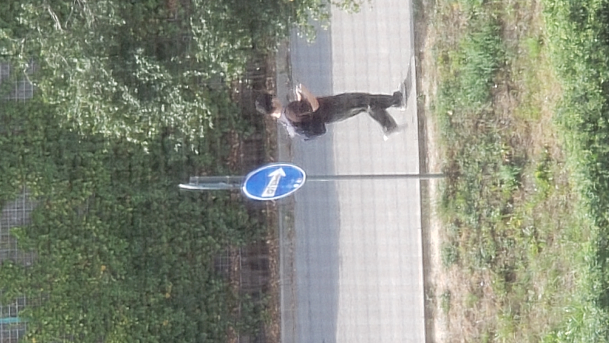 A lone person is walking on a sidewalk next to a street, facing right. They are wearing a dark t-shirt, dark pants, and white shoes. They have a backpack on their shoulders and are looking down at a mobile device in their hands. A blue, oval-shaped traffic sign with a white arrow pointing to the right is mounted on a metal pole beside the sidewalk. The background consists of a grassy embankment and a wall covered in green foliage, suggesting an outdoor urban or suburban setting. The lighting indicates it is daytime. This scene is in Prešov, Slovakia, near the Šariš Brewery.