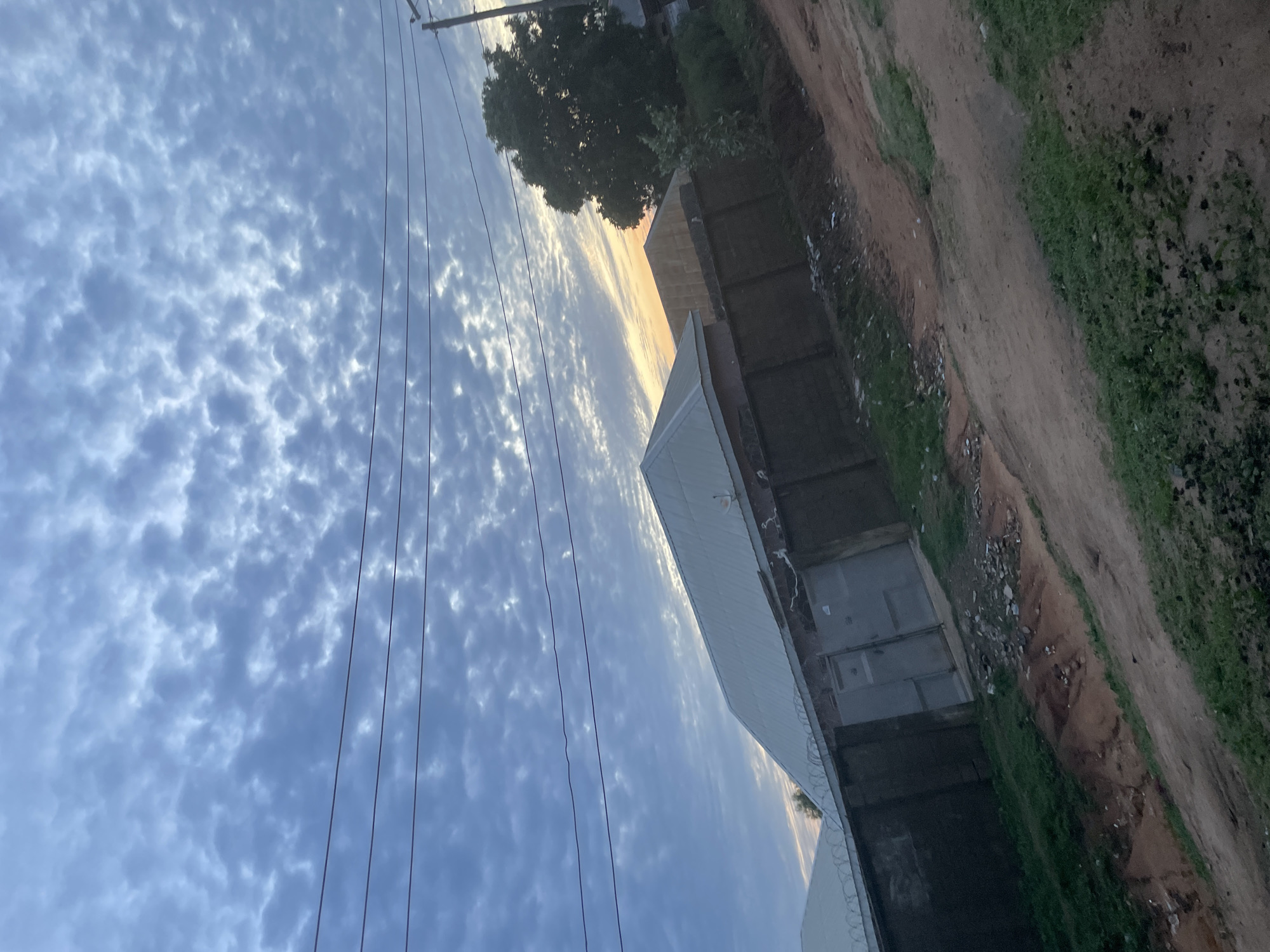 The image shows a street scene in Jalingo, Nigeria. The sky is filled with scattered clouds and a hint of sunset colors in the distance. Power lines run horizontally across the frame. In the foreground, a dirt road with patches of green grass leads up to a dark fence. Behind the fence, there are buildings with corrugated metal roofs and a large grey gate. A large tree is visible on the right side of the image, casting a shadow. Debris is scattered along the fence line and on the ground. The overall impression is a residential area at dusk.