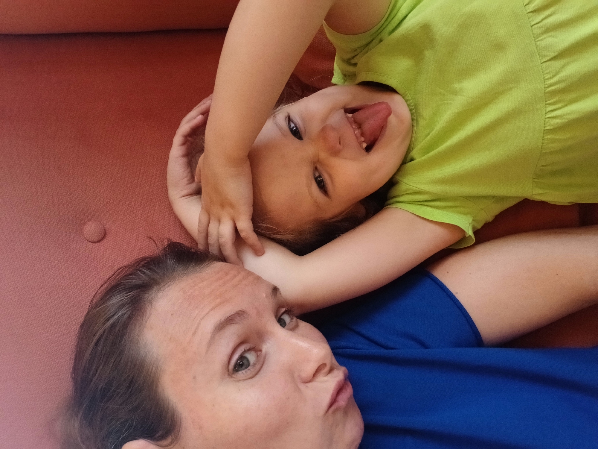 A woman and a young child are taking a selfie on a reddish-orange sofa. The woman, positioned on the left, is wearing a blue t-shirt and has her head tilted towards the camera with a pouting expression. The child, on the right, is wearing a light green dress and is sticking out their tongue while holding their hands on their head. The background is the textured surface of the sofa. The image does not contain any information to determine its location in Yekaterinburg, Russia.