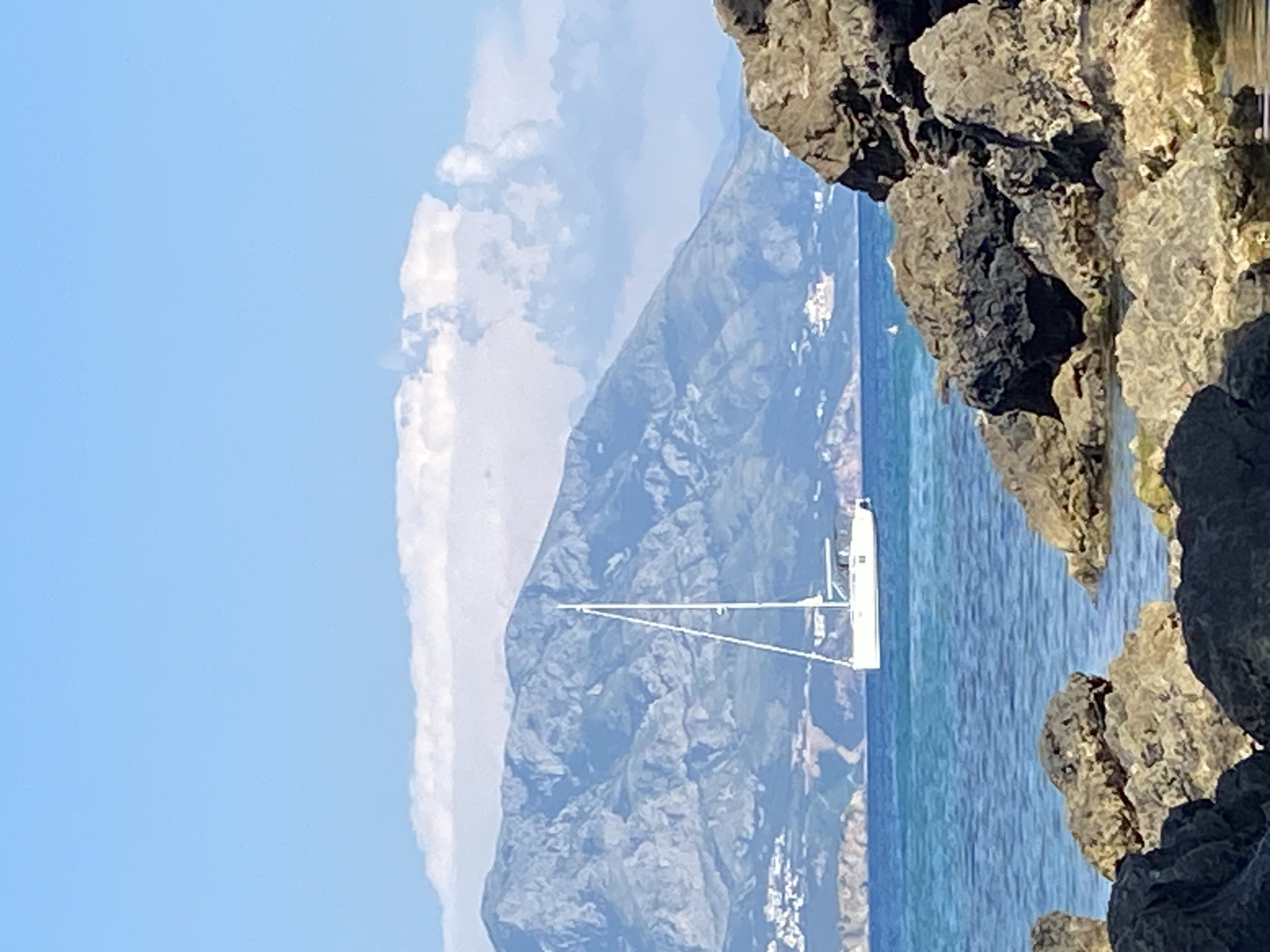 A sailboat is visible on the turquoise waters of the Adriatic Sea, with the mountainous landscape of Prijevor, Montenegro serving as a backdrop. In the foreground, a cluster of dark, rocky outcrops partially obscures the view of the sea and the sailboat. The sky is a clear blue, with a few wispy white clouds scattered near the horizon. The sailboat is white with a tall mast and sails, and appears to be stationary or moving slowly. Buildings are scattered on the slopes of the mountains, adding a touch of human presence to the otherwise natural scene.