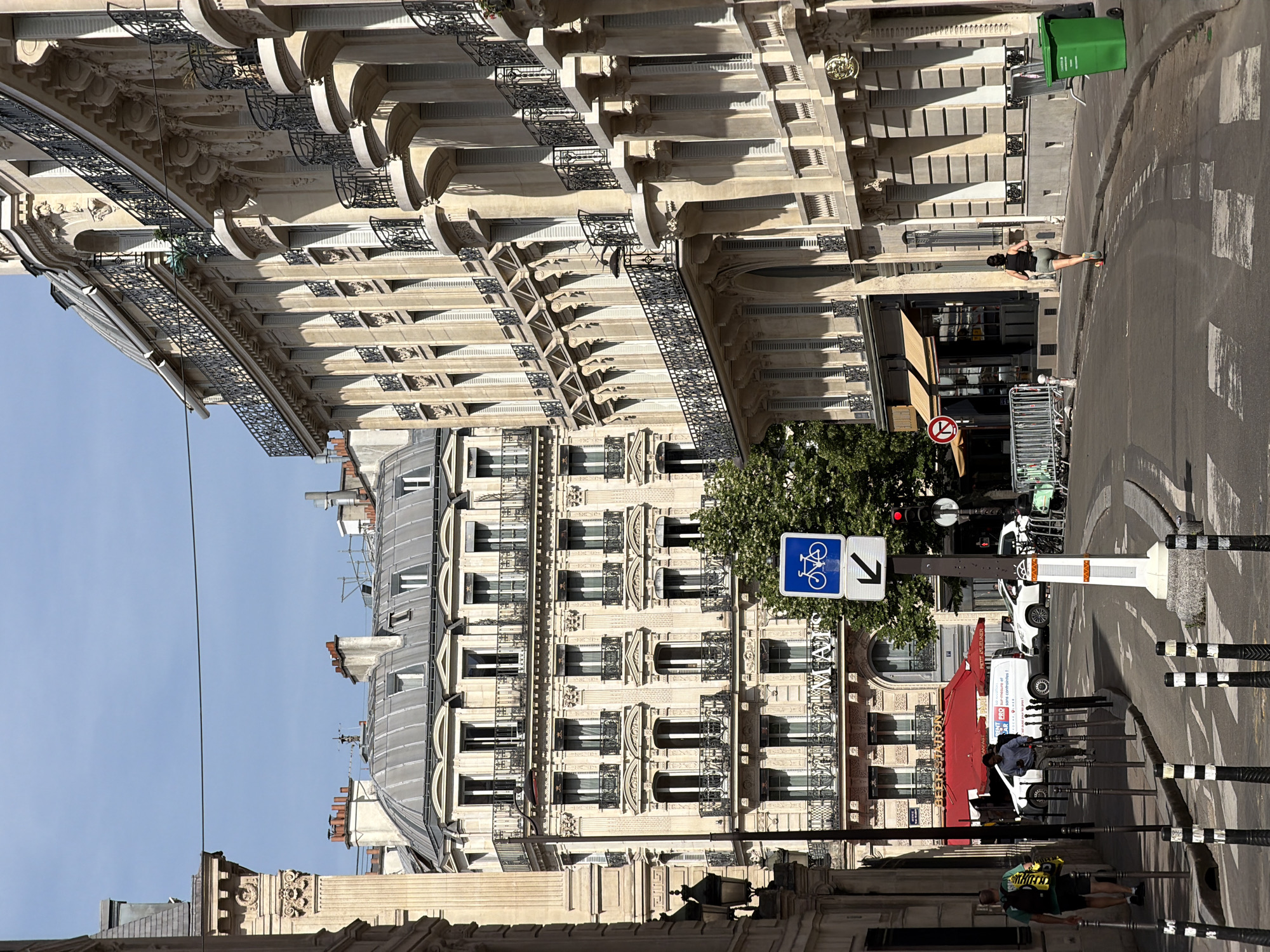 The image depicts a street in Paris, France, lined with ornate buildings featuring balconies and intricate architectural details. The sky is clear and blue. Street signs are visible, including one indicating a bicycle route. People are present on the street, one appearing to be walking and others near a "BEERSTATION" storefront. A van is parked nearby. Street furniture, such as bollards, and trash bins are also visible.