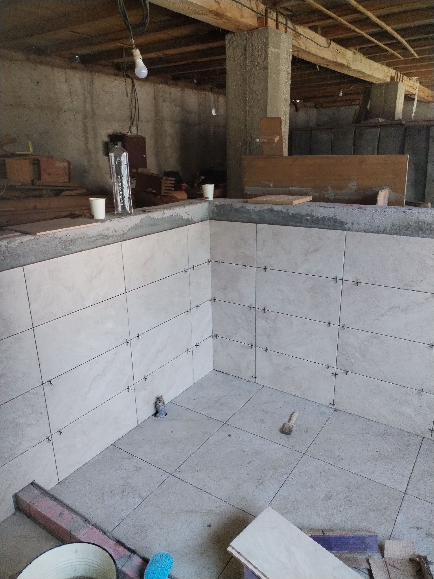 The image depicts the interior of a partially constructed building, possibly a workshop or unfinished room, with exposed wooden beams on the ceiling and concrete walls. The foreground shows a corner where the floor and walls are tiled with large, light-colored tiles. The tiling is in progress, with tile spacers visible between the tiles on the walls and floor. A small brush with wood shavings is resting on the tiled floor. In the background, there are stacks of lumber, construction materials, and some basic lighting fixtures. Two white paper cups are placed on the tiled ledge of the wall. A long, clear glass bottle with markings is also visible on the ledge. The overall impression is a scene of ongoing construction or renovation. The specific location in Lipănescu, Romania cannot be determined from the visual information provided in the image.