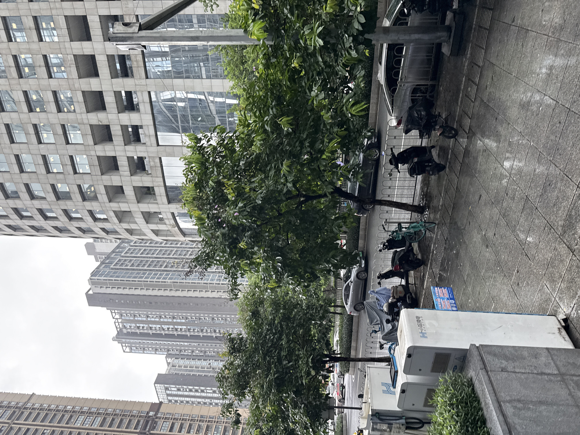 In Tianhe District, China, a city street scene unfolds under an overcast sky, with wet pavement indicating recent rain. The foreground features a sidewalk with several electric scooters and bicycles parked. Two white charging stations for electric scooters are prominent on the left, one with Chinese characters reading "H-小桔快电" (H-Jú Kuài Diàn). The sidewalk is paved with gray square tiles, and a metal grate runs along the edge.
Beyond the sidewalk, a street lined with trees separates the pedestrian area from vehicle traffic. Several cars, including a silver sedan and a dark-colored sedan, are visible on the road. The background is dominated by tall modern buildings. On the right, a large, cylindrical building with a grid-like facade of windows and stone panels stands prominently. To the left of this structure, a series of high-rise apartment buildings with repeating window patterns are visible. The overall impression is a busy urban environment with a mix of residential and commercial architecture.