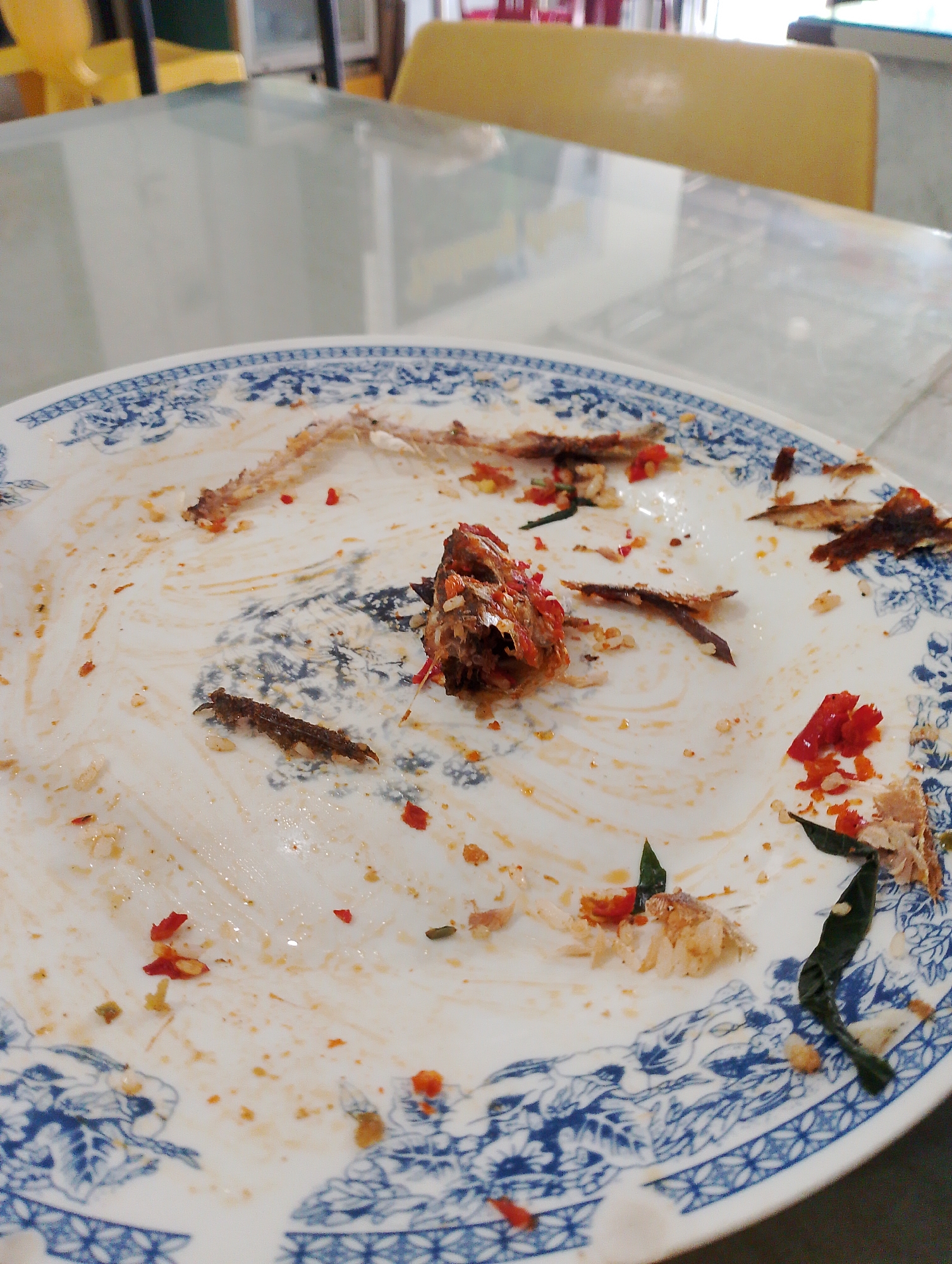 A close-up, eye-level shot shows a dirty white plate with a blue oriental pattern on the rim, resting on a reflective surface, likely a table. The plate is littered with the remnants of a meal, primarily consisting of fish bones, scattered pieces of red chili, and small flecks of white rice. A larger piece of cooked fish, appearing to be the head, is visible near the center of the plate. The background is blurred, showing a hint of yellow seating and a glass-topped table, suggesting an indoor dining environment, possibly a restaurant. The lighting is somewhat dim, creating reflections on the table surface.
