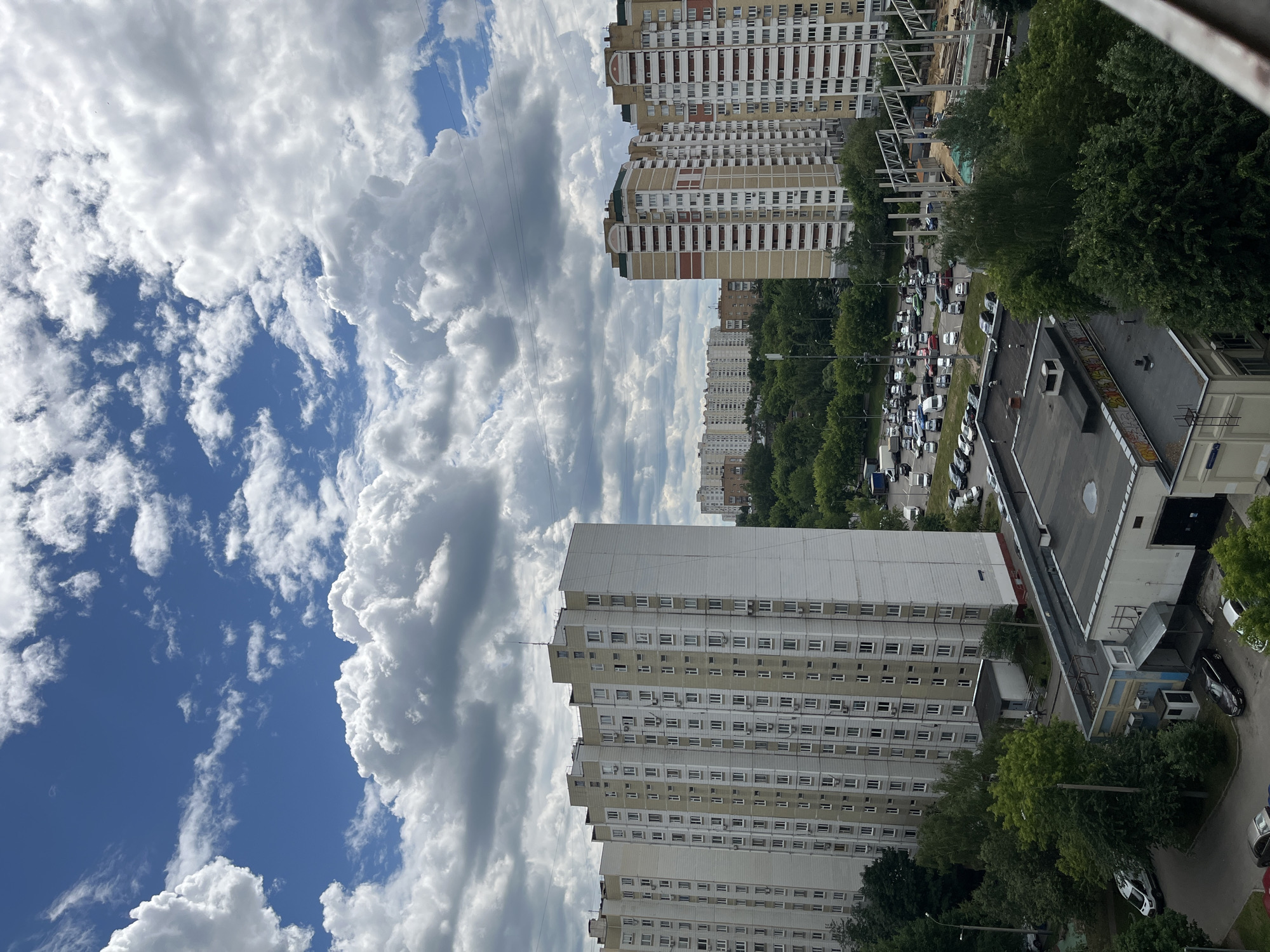 The image shows a wide shot of a residential area in Moscow, Russia. The foreground features a low-rise building with a gray roof, partially obscured by trees. In the middle ground, a large, multi-story apartment building with a white and beige facade dominates the left side of the frame. To the right of this building, another tall apartment building with a beige and red facade is visible, along with a construction site featuring a steel framework. A parking lot filled with numerous cars is situated between the buildings and the greenery. The background consists of more apartment buildings and dense trees, suggesting a suburban or mixed-use urban environment. The sky above is a vibrant blue, dotted with large, fluffy white clouds, indicating fair weather. Power lines are visible crisscrossing the sky. No people are clearly discernible in the image.