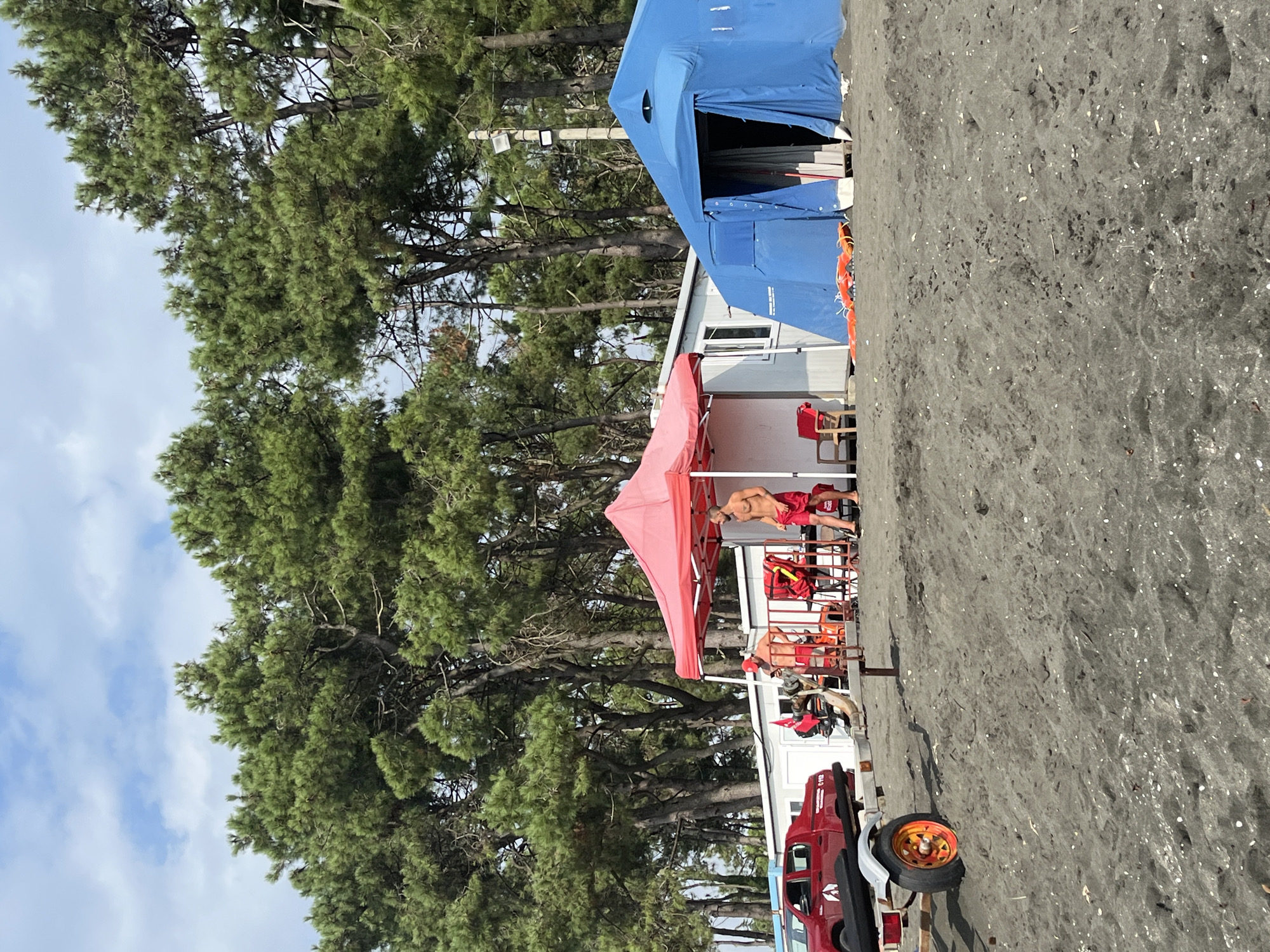 This image depicts a beach scene in Ureki, Georgia, characterized by dark sand and a backdrop of lush pine trees under a partially cloudy sky. In the foreground, the dark, wet sand is scattered with small white shells and debris. To the left, a red pickup truck is parked next to a trailer with a visible orange wheel.
Further back, several structures are present. A white building with windows is partially visible. A red pop-up tent is set up in front of it. Adjacent to the red tent is a large blue tent. Several people are visible near the structures. Two men in red shorts and one in a red hat are standing and interacting. One man appears to be holding a phone. Lifeguard chairs and equipment, including orange flotation devices, are also present. The overall atmosphere suggests a functional beach area, likely a lifeguard station or an operational base for beach activities. The presence of the truck, trailer, and tents indicates a setup for either a business or a service.