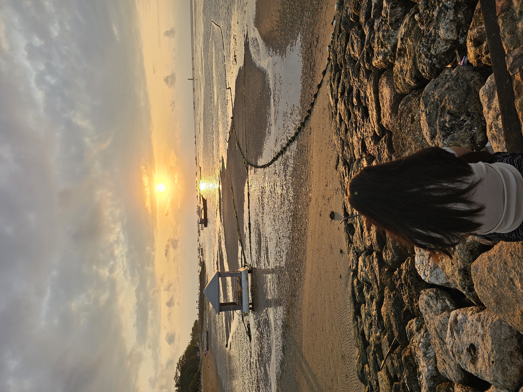 The image depicts a serene sunset scene at Sanur Beach, Denpasar, Indonesia. In the foreground, a person with long, dark hair is seated on a pile of large, rough rocks, facing away from the camera. The person is wearing a light-colored top and patterned bottoms. The rocks lead down to a sandy beach area where a small, open-sided shelter on a platform is situated in shallow water. The water recedes to reveal wet sand and small pools, reflecting the warm glow of the setting sun. Further out at sea, several boats are visible, some anchored and others appearing to be moving. A thin, dark line, possibly a fishing net or marker rope, extends across the water in the mid-ground. The sky is filled with dramatic clouds, tinged with orange and yellow hues from the sun, which is low on the horizon. In the far distance, a lighthouse or beacon can be seen. The overall atmosphere is tranquil and picturesque, capturing the beauty of a coastal sunset in Bali.