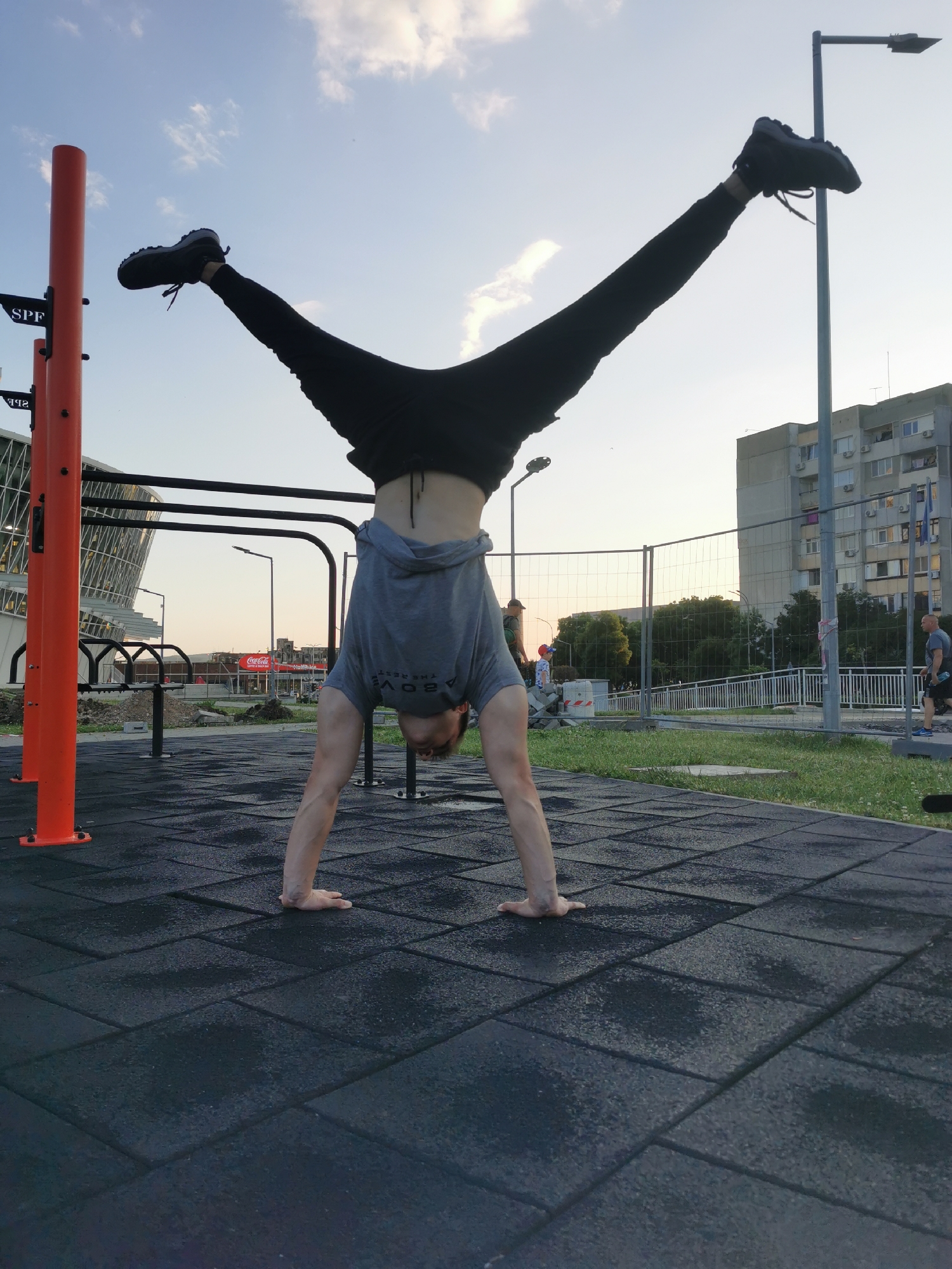 A person is performing a handstand with legs spread in a V-shape at an outdoor fitness park in Burgas, Bulgaria. The individual is wearing black pants, a gray t-shirt, and black sneakers. Their hands are planted on a dark, rubberized surface. Behind the person are various pieces of fitness equipment, including orange vertical posts and black horizontal bars.
In the background, there is a modern stadium with a glass façade and a building with numerous windows and air conditioning units, situated near a metal chain link fence with trees behind it. A few people are walking in the background near the building. The sky is light blue with some clouds. The location is identifiable as the fitness park near the Lazur Stadium in Burgas.