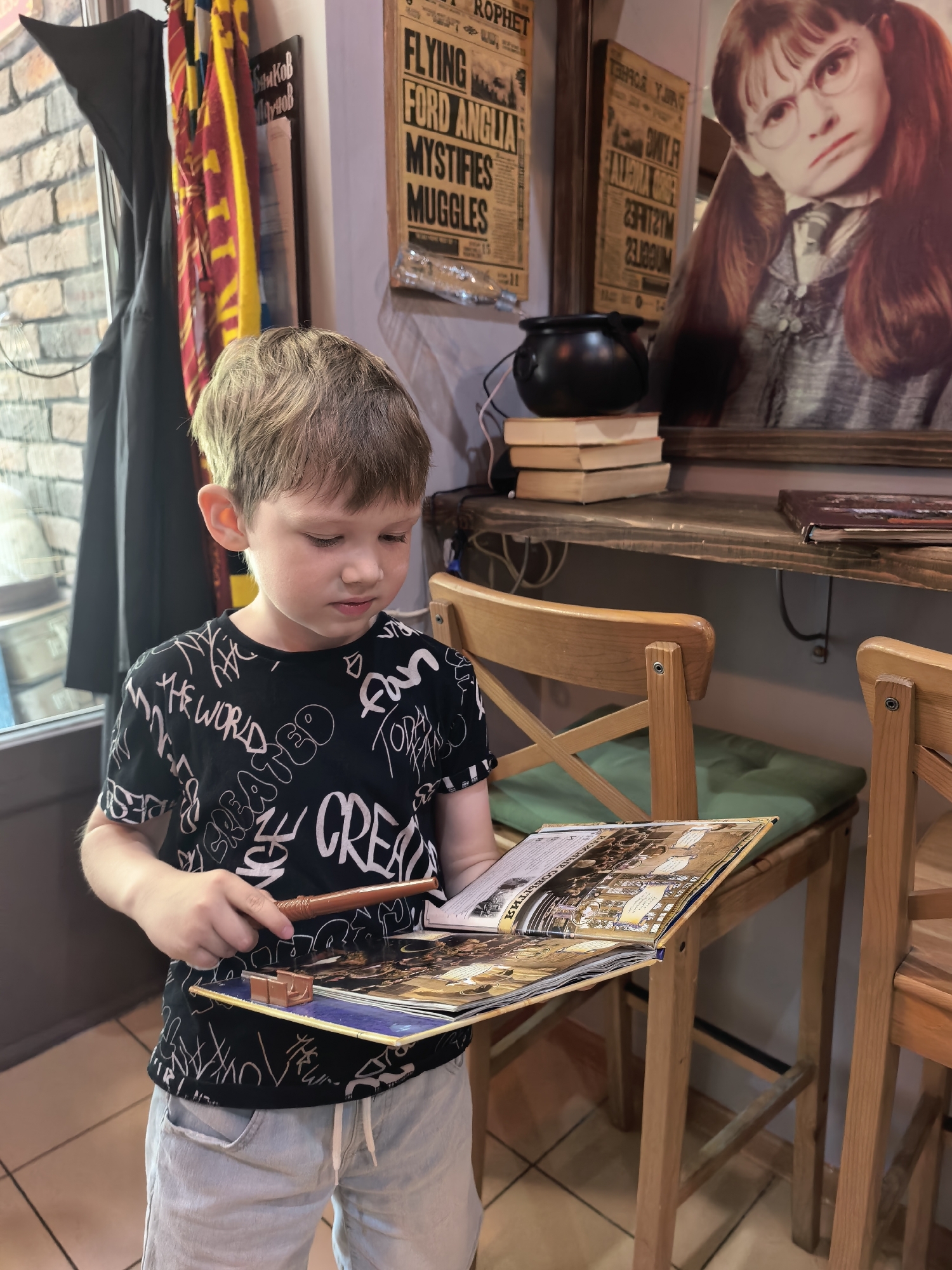 A young boy with light brown hair, wearing a black t-shirt with white lettering and light blue shorts, stands in a room that appears to be themed around Harry Potter. He is holding an open book or magazine in his hands and a small, brown wand. The book is open to a page with colorful illustrations. In the background, there are several Harry Potter-themed decorations, including a black cauldron, stacks of books, and posters or images of characters from the series. A large cutout of a character with red hair and glasses is visible on the right. Hanging on the left side of the image are what appear to be Hogwarts house scarves. The setting seems to be an indoor establishment, possibly a cafe or shop, with wooden chairs and tables visible. The overall impression is of a child engrossed in a magical world. The text "FLYING FORD ANGLIA", "MYSTIFIES", "MUGGLES", and "CREATED THE WORLD" is visible on the posters and the boy's shirt. The location is Sterlitamak, Russia.