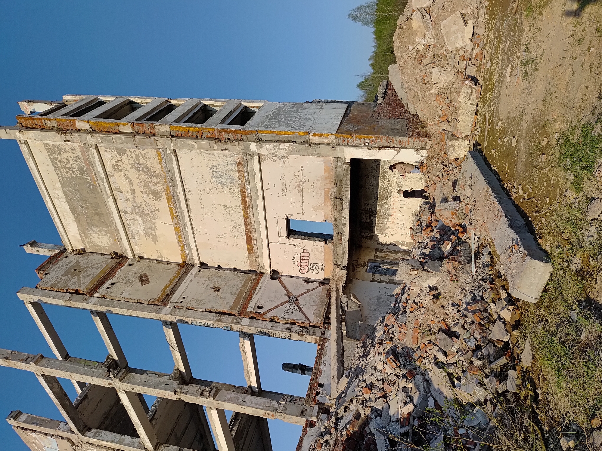 The image depicts the ruins of a building in Марковский, Russia. The remnants of the structure consist of concrete and brick walls, with several window openings. Sections of the building appear to have collapsed, leaving piles of rubble. Two figures are visible inside the building. The sky is clear. Vegetation, including trees and grass, is present in the surrounding area. Graffiti is visible on the interior walls of the building.