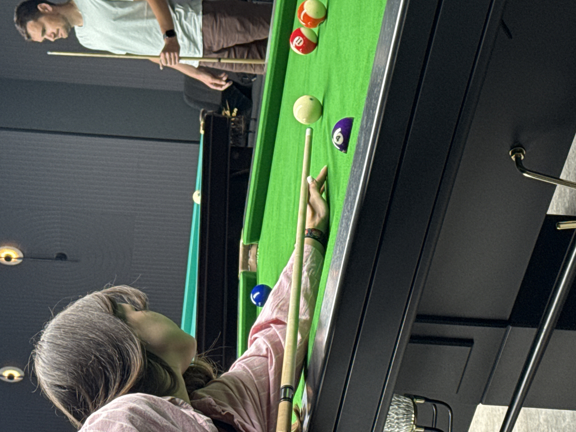 In a dimly lit indoor room, a woman with long brown hair, wearing a pink shirt, is positioned to play pool. She is hunched over a green felt pool table, with her right hand resting on the cue stick, poised to strike a ball. A cue ball is visible in front of her, and other pool balls, including a purple ball with the number 9, and two numbered red and orange balls, are scattered on the table. In the background, a man wearing a light blue t-shirt and brown shorts stands holding a cue stick, observing the game. The room features a dark gray wall and a ceiling with circular lights. The image does not provide specific details to determine the location in the city of Balaklavsky District, Ukraine.