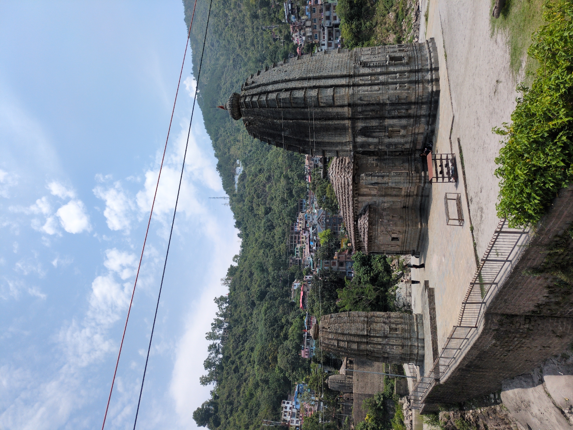 The image depicts the historic Triloknath Temple in the city of Mandi, India, nestled within the Himalayas. The temple complex features several ancient stone structures with intricate carvings. The main temple, a tall, shikhara-style structure, is prominently displayed, characterized by its tiered design and stone ornamentation. To the left, a smaller, similarly constructed temple stands, partially obscured. The ground area in front of the temples is paved with stone, and a metal railing lines the perimeter, overlooking a rocky, dry riverbed. A few individuals are present, two standing near the smaller temple and one seated near the main temple structure. Behind the temple complex, a dense green hillside rises, dotted with buildings of varying colors and architectural styles, indicating the presence of a town or city. Power lines traverse the upper portion of the frame, against a backdrop of a bright blue sky with scattered white clouds.