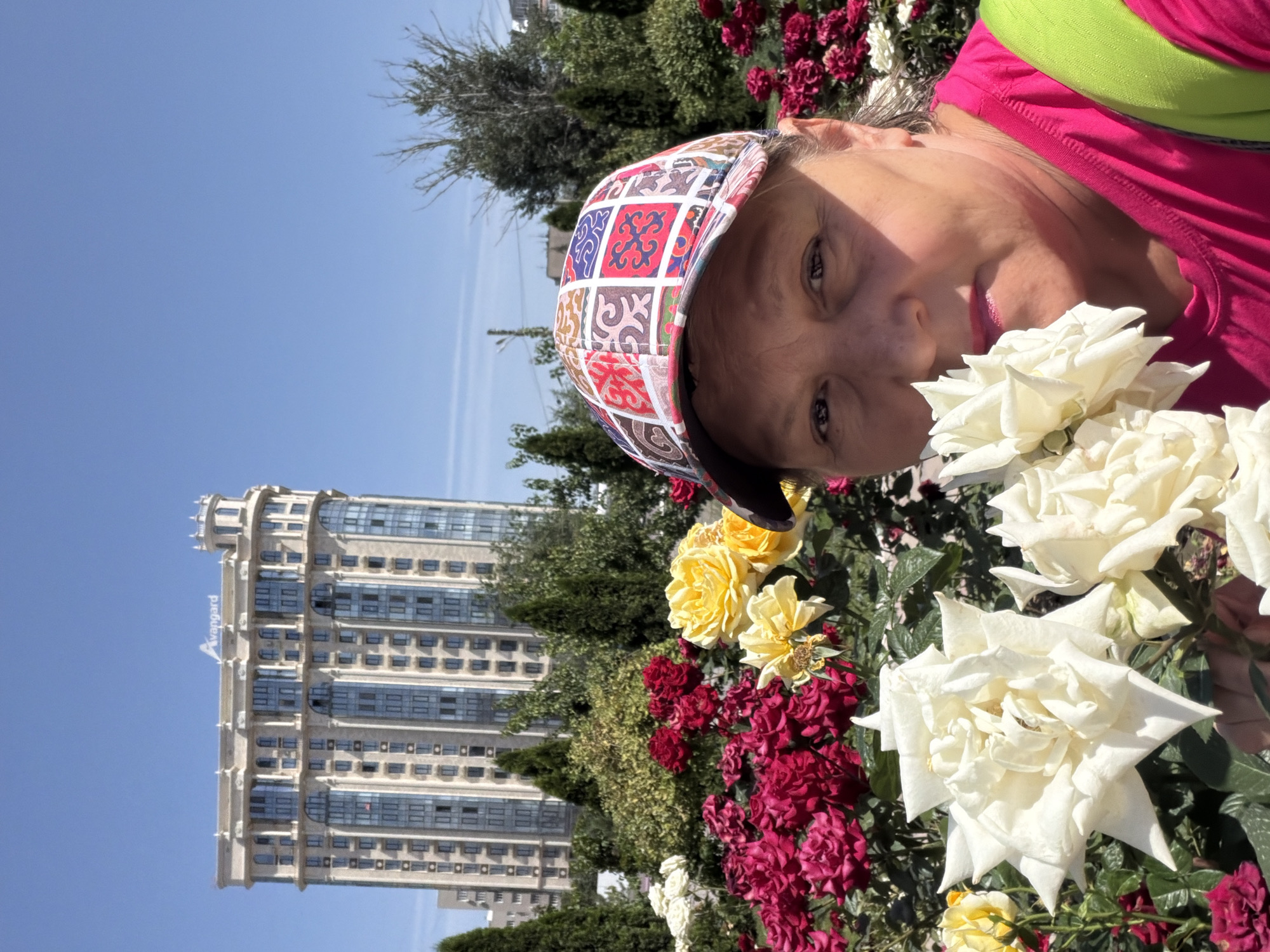 In an outdoor scene, a woman wearing a brightly colored baseball cap and a pink t-shirt is taking a selfie. She is positioned in the foreground, partially obscuring a vibrant rose garden filled with red, yellow, and white roses. Behind the garden, a tall, ornate building with many windows stands against a clear blue sky. The building appears to be an apartment complex or hotel, with decorative architectural elements. The overall setting suggests a park or public space in a city.