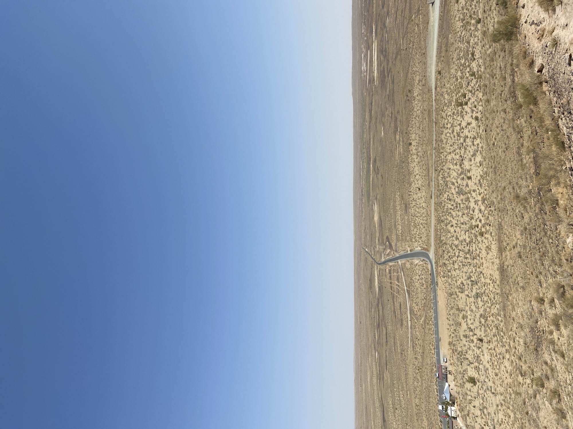 The image shows a vast, arid desert landscape under a clear blue sky. A paved road cuts through the barren terrain, stretching towards the horizon. On the left side of the road, a small cluster of buildings, possibly a rest stop or small settlement, is visible. Several white vehicles are parked near these structures. The ground is covered with dry, sparse vegetation and appears uneven, with some rocky outcrops in the foreground. The horizon is flat, with a hazy appearance in the distance, suggesting a wide, open expanse. The location is identified as the city of Beshtom, Uzbekistan, a region known for its desert climate. There are no discernible people in the image.
