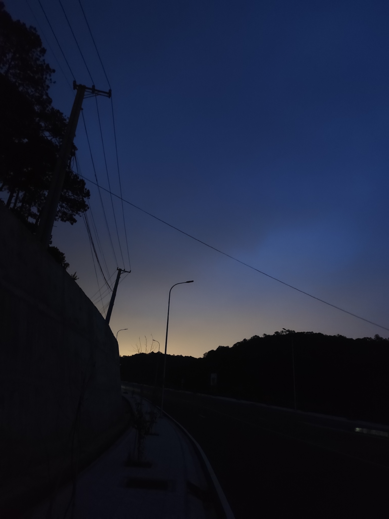A low-angle shot depicts a road and adjacent wall at dusk in what appears to be the city of Da Lat, Vietnam. A thick concrete wall looms on the left, contrasting with the dark silhouette of a tree and a utility pole with exposed wiring. Beyond the wall, streetlights dimly illuminate a road, which is mostly shrouded in darkness. Across the road, a hill covered with trees forms a dark skyline against the faint glow of the setting sun. The sky transitions from a light golden hue near the horizon to a deep blue at the top of the frame.