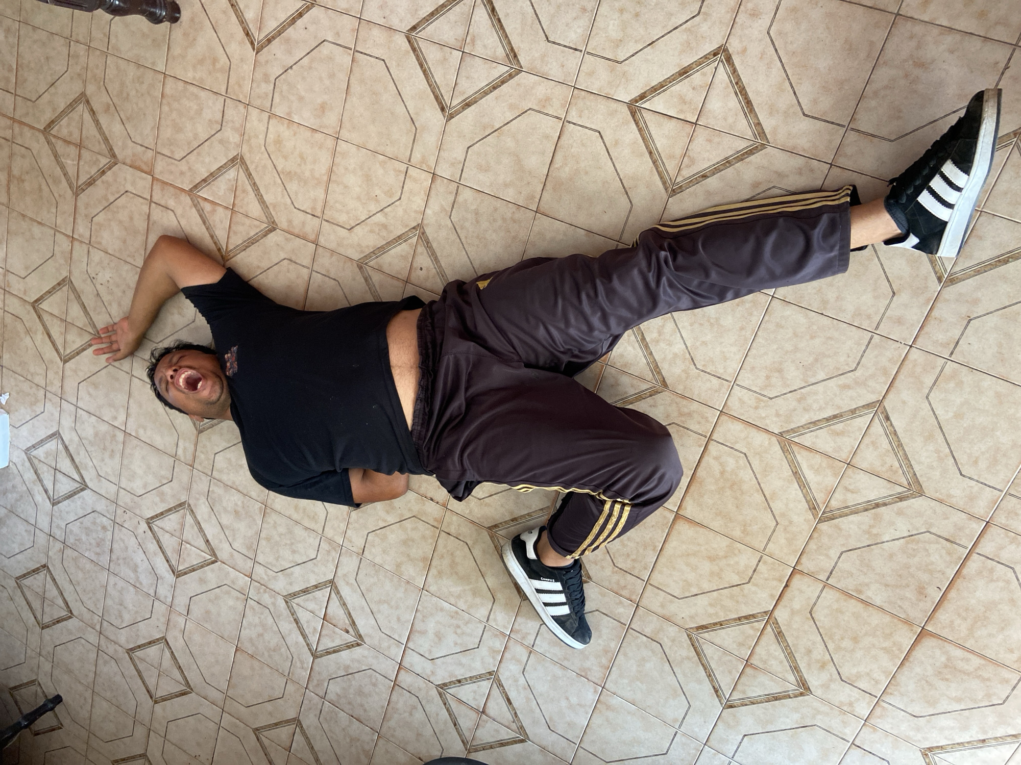A man lies prone on a tiled floor. The man is wearing black shorts and a black T-shirt that is slightly raised, revealing his stomach. He is also wearing gray track pants with gold stripes down the sides and black and white Adidas sneakers. His mouth is open in a yell. The tiled floor has a geometric pattern.
Tinaquillo, Venezuela is NOT mentioned in the picture.