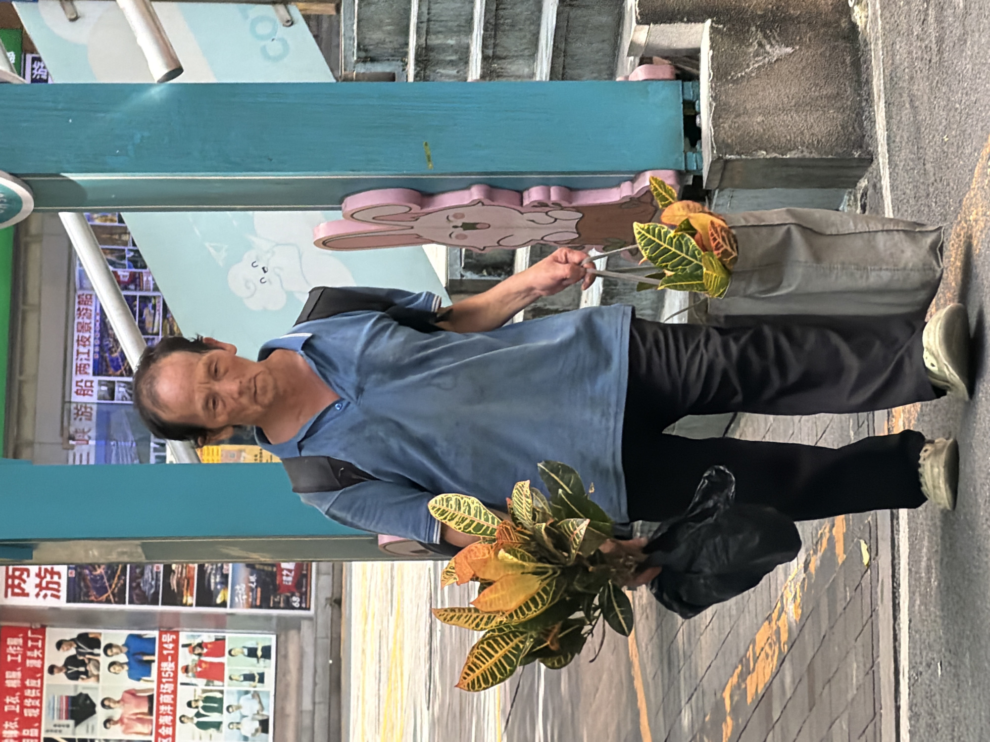 The image depicts a man walking on a sidewalk in Yuzhong District, China. He is carrying a bouquet of colorful plants in his left hand, wrapped in a black plastic bag, and a grey tote bag containing more plants in his right hand. The man is wearing a light blue polo shirt, dark pants, and light-colored shoes. He has a backpack on his shoulders. Behind him, a turquoise-colored pillar with a pink cartoon rabbit decoration is visible, as well as a staircase. Advertisements are displayed on a wall to the left of the man. The overall environment suggests an urban setting.