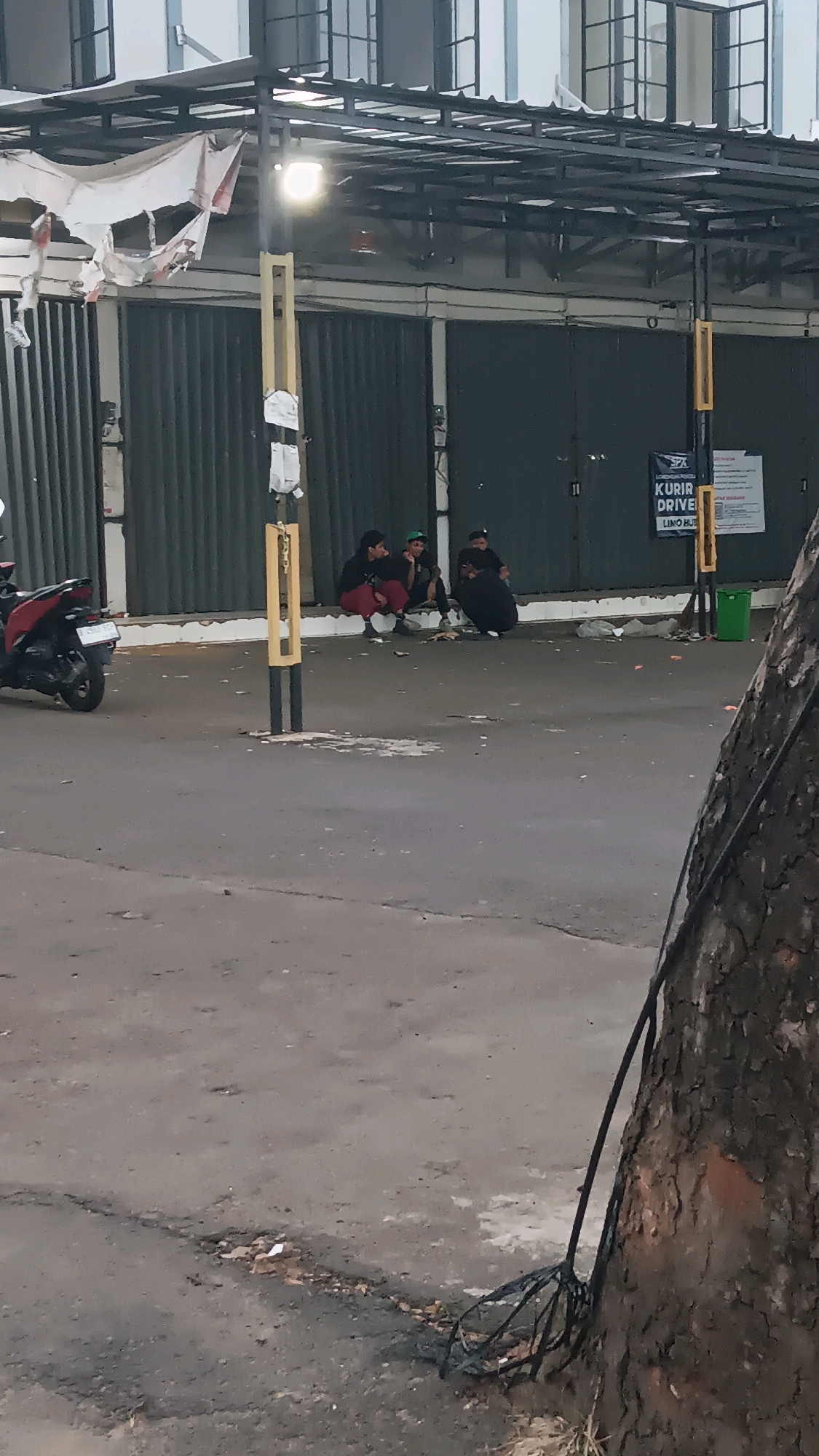 Three individuals are crouched on the pavement in front of a corrugated metal facade. The individual on the left wears a red top and dark pants, the middle person wears a dark top and dark pants with a green cap, and the person on the right is dressed in all black. A red motorcycle is parked to the left of the frame. The background consists of a building with multiple windows and a covered walkway. A sign for "SPX KURIR DRIVE" is visible on the wall. A green trash can is situated to the right. The scene appears to be outdoors, possibly on a street or in front of a commercial establishment in Depok, Indonesia. The ground is paved and shows signs of wear and debris. A tree trunk with rough bark is prominent in the right foreground.