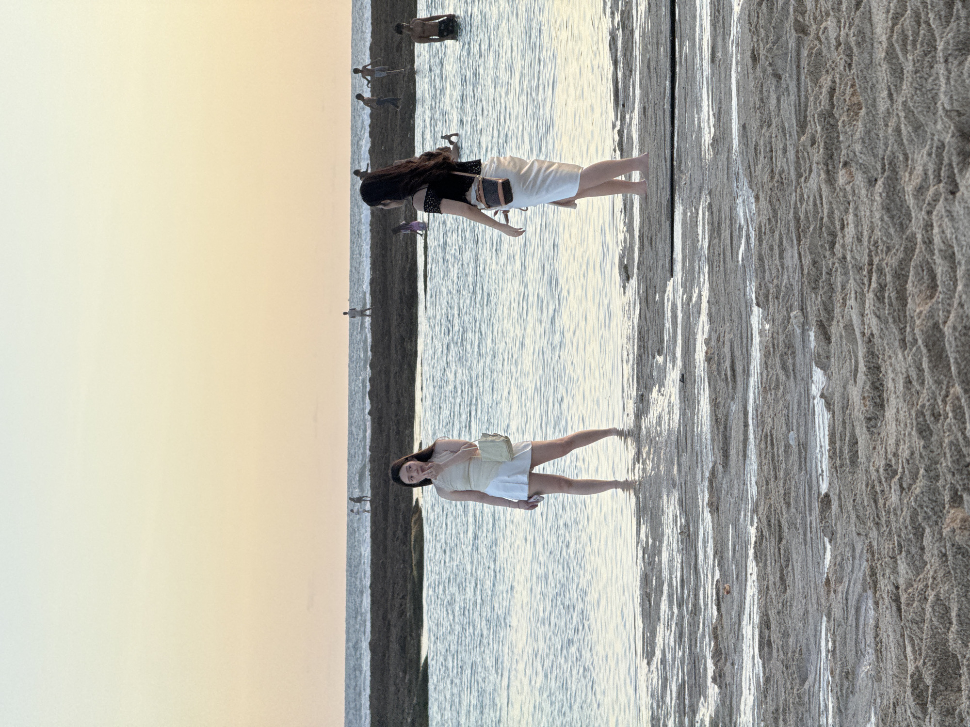 Two women stand in shallow water at the edge of a beach in Kuta Selatan, Indonesia. The woman closest to the camera wears a light-colored dress and carries a purse, while the other woman is dressed in a dark top and a white skirt with a purse. In the background, several people are wading further into the ocean. The beach sand is visible on the right side of the frame, and the sky is a gradient of light yellow and orange.