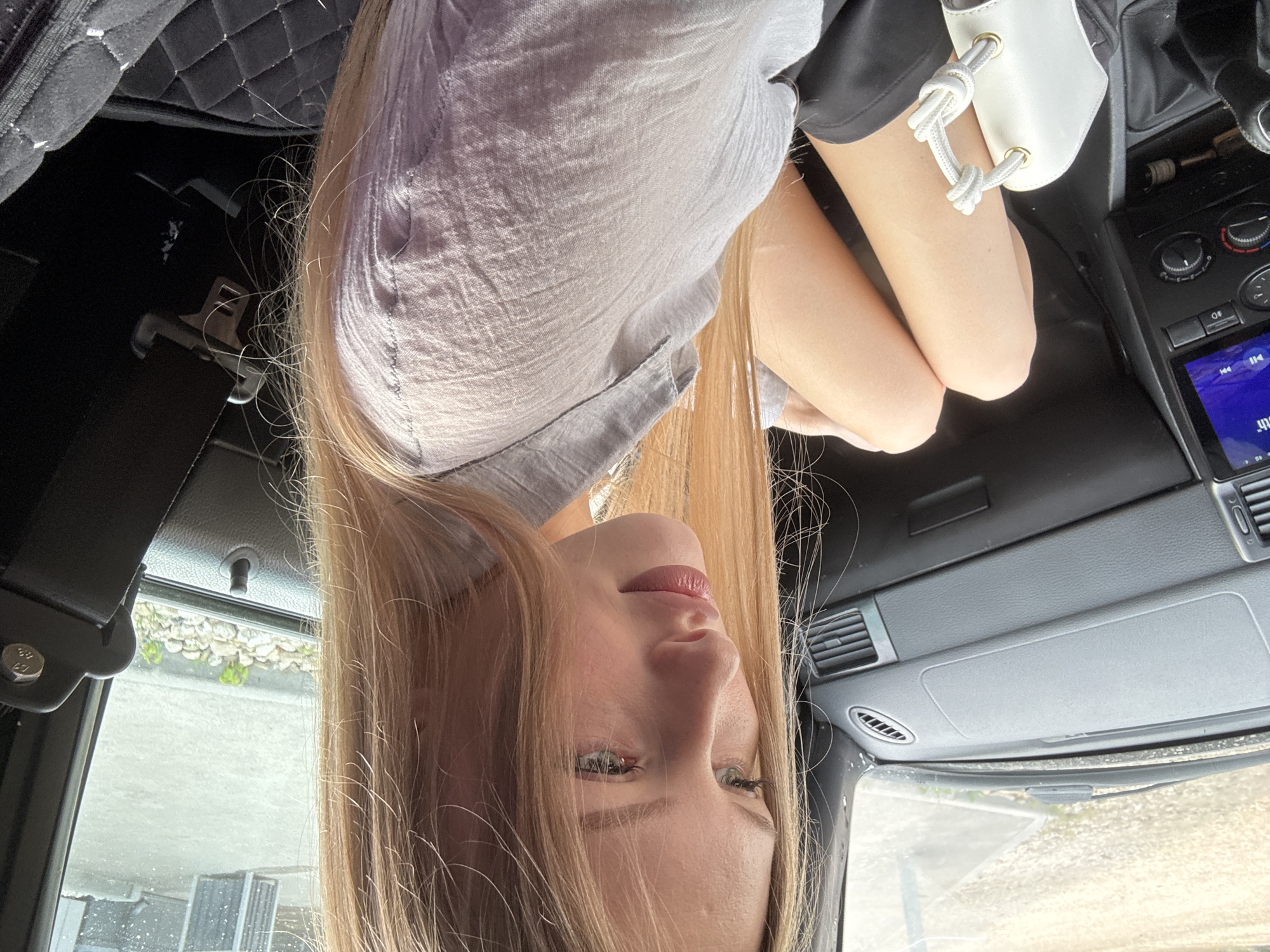 A young woman with long blonde hair sits in the passenger seat of a car. She is wearing a light grey, slightly sheer collared shirt and black shorts. Her legs are crossed, and she holds a small white purse with a knotted rope handle. The car's interior is visible, including the dashboard, a gear shift, and a car seatbelt. The woman looks away from the camera, her head tilted downwards. The environment outside the car appears to be a paved area with some gravel and small plants, suggestive of an urban or suburban setting.