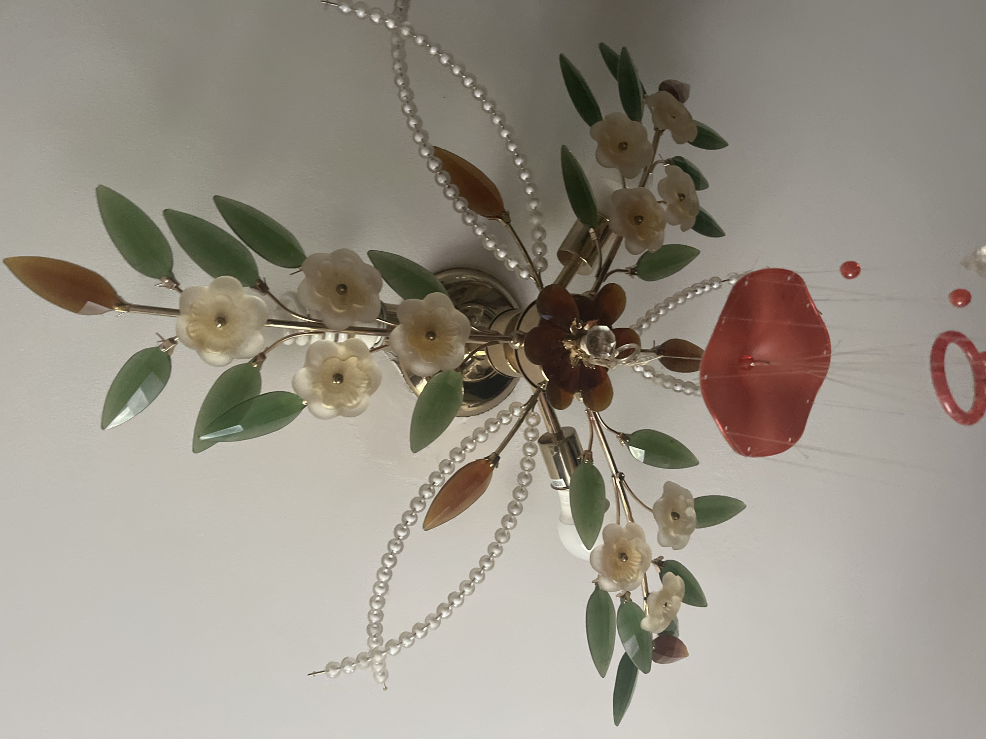 This is a close-up, eye-level, indoor shot of a decorative ceiling light fixture. The fixture is gold-toned and features a floral motif with multiple tiers of faux flowers, green jade-like leaves, and amber-colored leaf-shaped accents. Strands of faux pearls are draped around the fixture, and a red, petal-shaped ornament hangs from the bottom, adorned with thin strings and small red beads. The background is a plain, light-colored wall, likely a ceiling. There are no people or animals visible in the image, and the setting is consistent with an interior space in a residential or commercial building. The location is unknown, but the request specifies Minsk, Belarus.