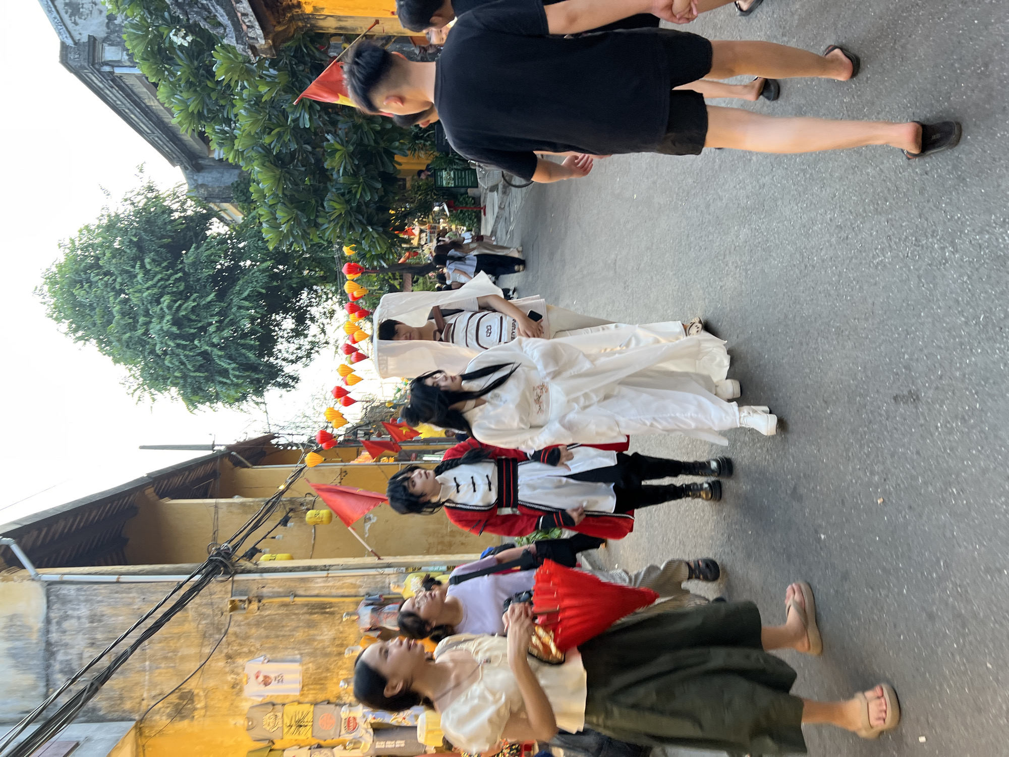 This is a vertical, full-body shot of a group of people in a street, likely in Hội An, Vietnam, given the characteristic yellow buildings and the presence of decorative lanterns. The time of day appears to be late afternoon or early evening, with soft, warm lighting.
The scene features several individuals. In the foreground, a woman in a light-colored top and a dark green skirt is holding a red fan. Next to her, another person in a white shirt and dark pants looks up. Further behind them, a person in a red and white outfit with black pants and shoes stands out. Above this person, two individuals are lying down or reclining on the street, one dressed in white and the other in white with a backpack. At the very top of the frame, a person in a black t-shirt and shorts walks away from the camera.
The activity seems to be some kind of performance or staged event, possibly a reenactment or a themed photoshoot, given the costumes and the poses of the people lying down. The presence of red and yellow lanterns strung across the street, along with small red flags, suggests a festive atmosphere. The background shows traditional architecture with muted colors and overhead electrical wires. There is no visible text in the image.