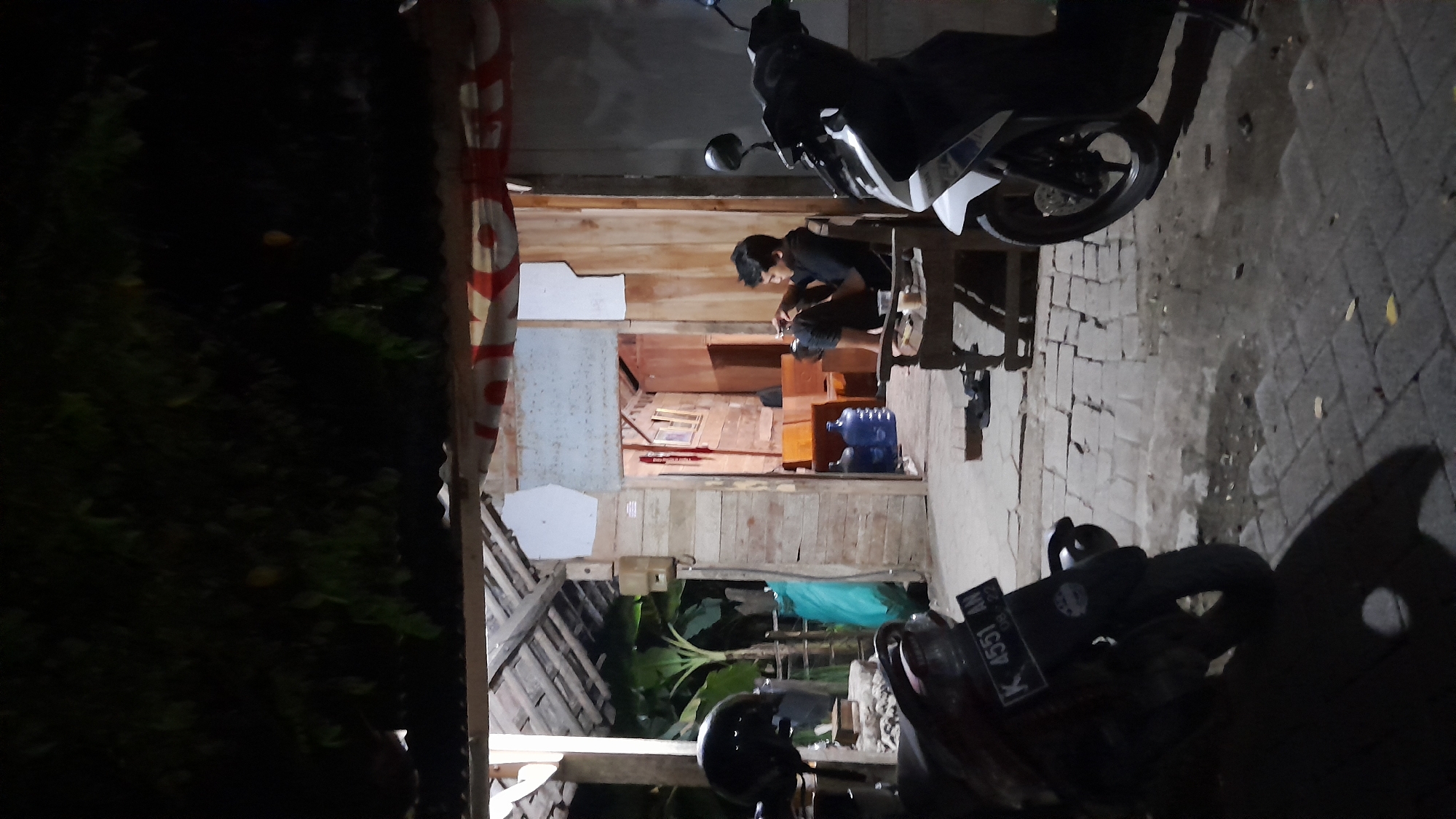 The image depicts an outdoor scene at night in Sumberarum, Indonesia. In the foreground, a dark-colored scooter with a license plate reading "K 4551 AN" is partially visible on the left, and a white and black scooter is parked on the right. A person wearing a black t-shirt is seated on a wooden chair in front of a wooden structure. The structure appears to be a shop or dwelling with an open entrance revealing interior details such as wooden walls and furniture. There are two large blue water jugs visible inside the structure. The ground is paved with brick-like stones. The overall lighting suggests it is nighttime, with artificial light illuminating the area.
