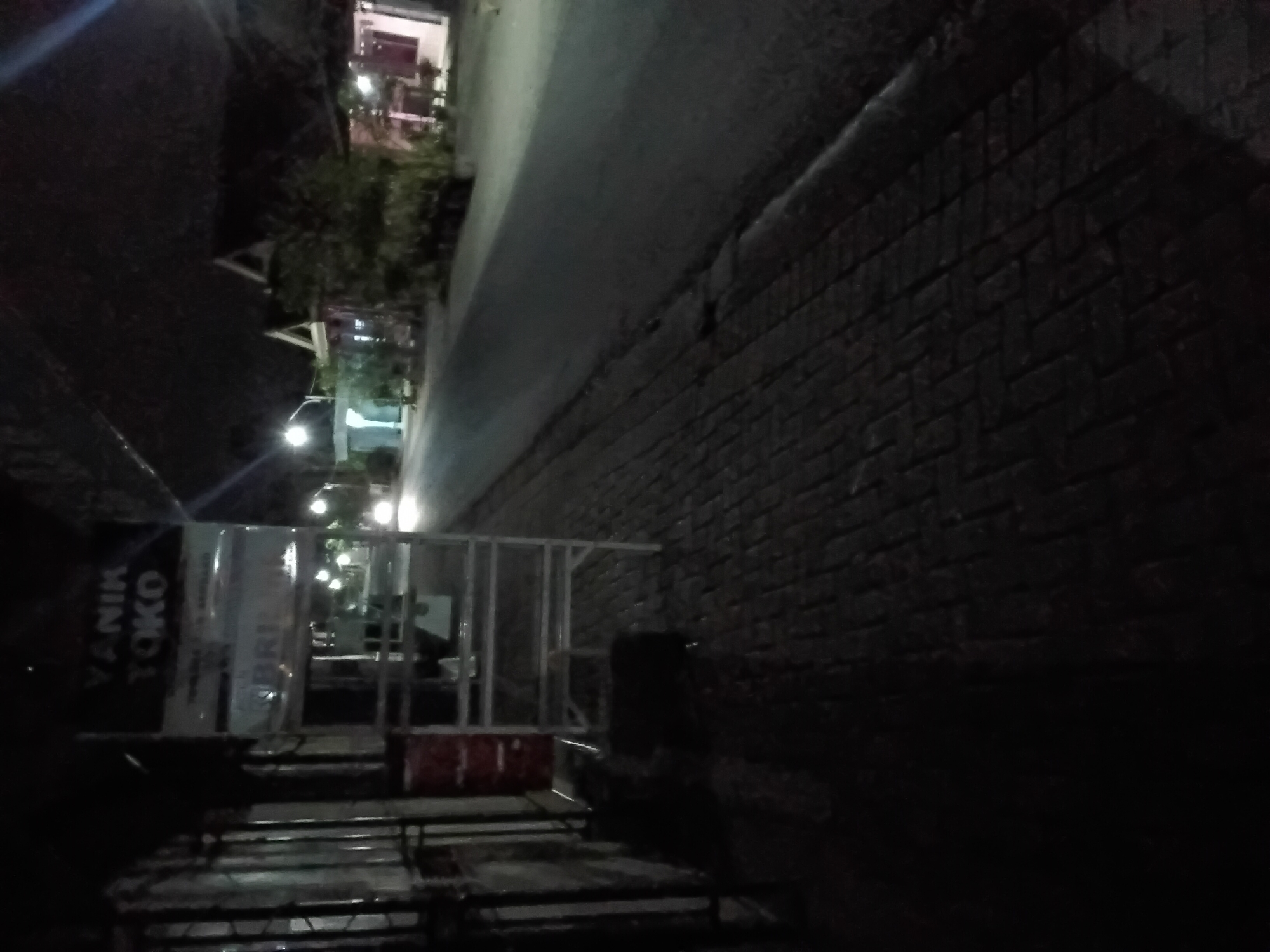 The image is a nighttime street scene in Trenggalek, Indonesia. On the left side of the frame, a metal stand with signs is visible, one of which reads "YANIK TOKO". Adjacent to the stand is a red barrel. The ground is paved with dark bricks. In the background, a road stretches into the distance, with streetlights illuminating the area. Several buildings with pitched roofs are visible on the right side of the road, with trees and vegetation in front of them. Headlights from a vehicle are visible on the road. The overall scene is dark and illuminated by artificial lights.