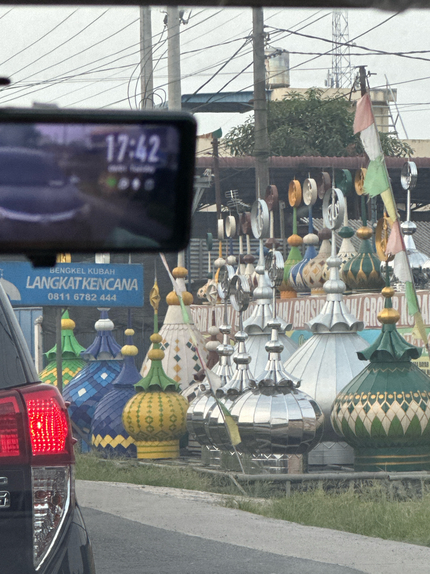 The image depicts a roadside view in the city of Binjai, Indonesia. In the foreground, the taillight of a black car is visible on the left, reflecting red light. The car is positioned on a paved road next to a grassy verge. Behind the car, a large number of dome-shaped structures, likely decorative elements for mosques or similar buildings, are displayed. These domes vary in color, including blue, green, yellow, and silver, and are adorned with various patterns. A blue sign with white text reads "BENGKEL KUBAH LANGKATKENCANA" and a phone number "0811 6782 444," indicating a business selling or manufacturing these domes. In the background, utility poles with numerous electrical wires are prominent, along with some buildings and trees. The sky is overcast, suggesting a cloudy day. The overall scene is of a commercial area specializing in religious architectural elements, situated along a road.