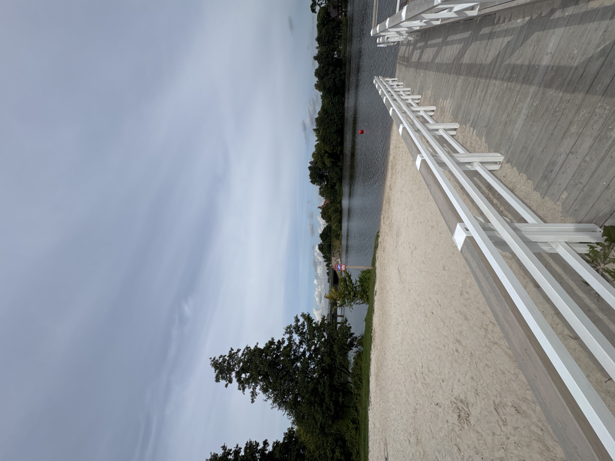 The image depicts a serene view of a sandy beach leading to a wooden pier with white railings, situated on the shore of a calm body of water in Sestroretsk, Russia. The sky is overcast with scattered clouds, and on the opposite bank of the water, dense green trees form a natural border. A bridge is visible in the distance, spanning the water, with some buildings nestled amongst the trees. A red buoy floats in the water near the pier. The scene suggests a quiet, recreational area.