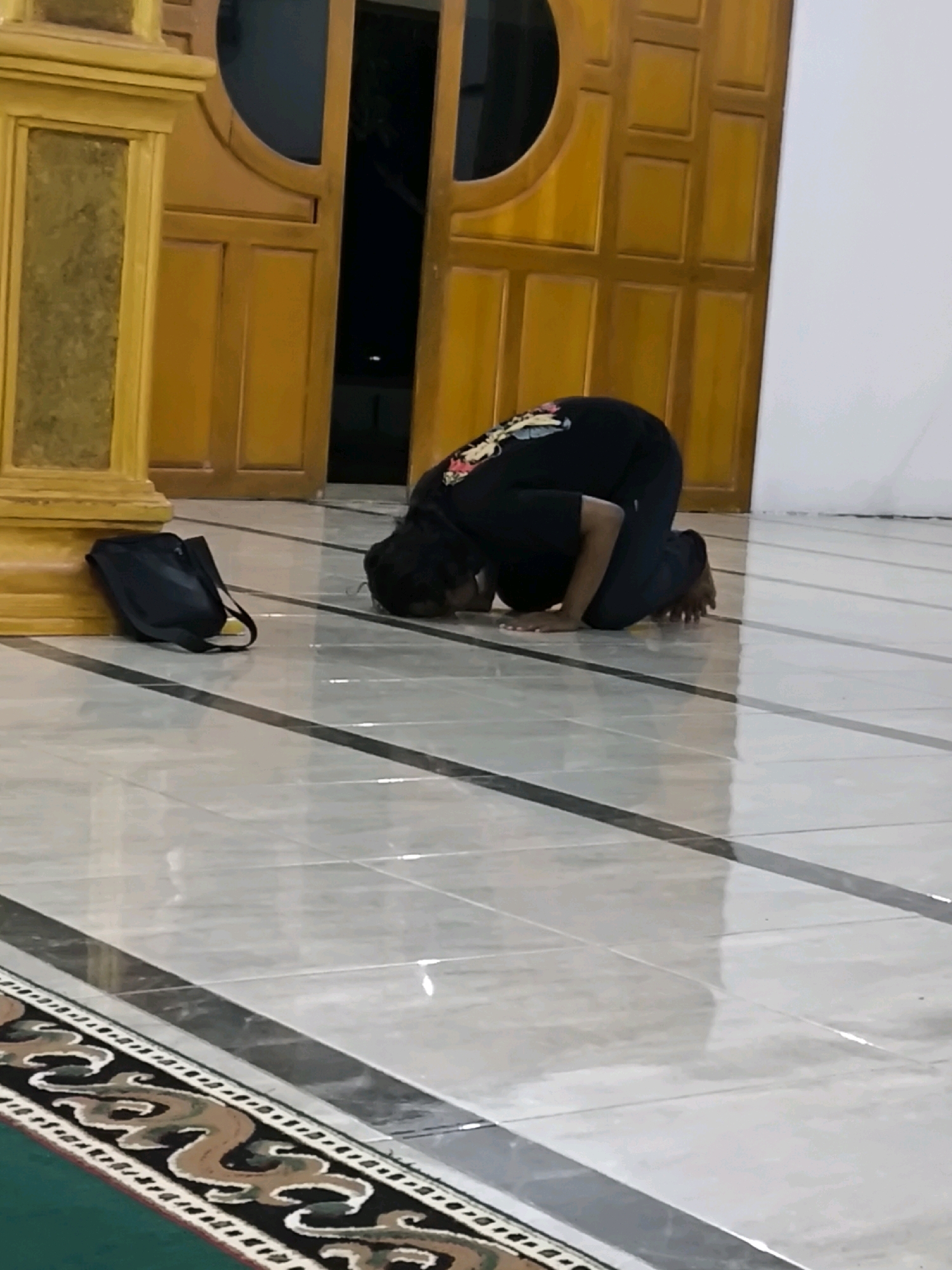 A person in a black shirt and pants is kneeling on a tiled floor, with their forehead touching the ground. They are facing away from the camera, toward a set of wooden double doors. A black bag is placed on the floor to the left of the person, leaning against a wooden pillar. The floor is tiled with a glossy finish and has dark grey lines dividing the tiles. A decorative border runs along the bottom of the image. The background shows the wooden doors and a white wall. The location is identified as Unknown, Indonesia.