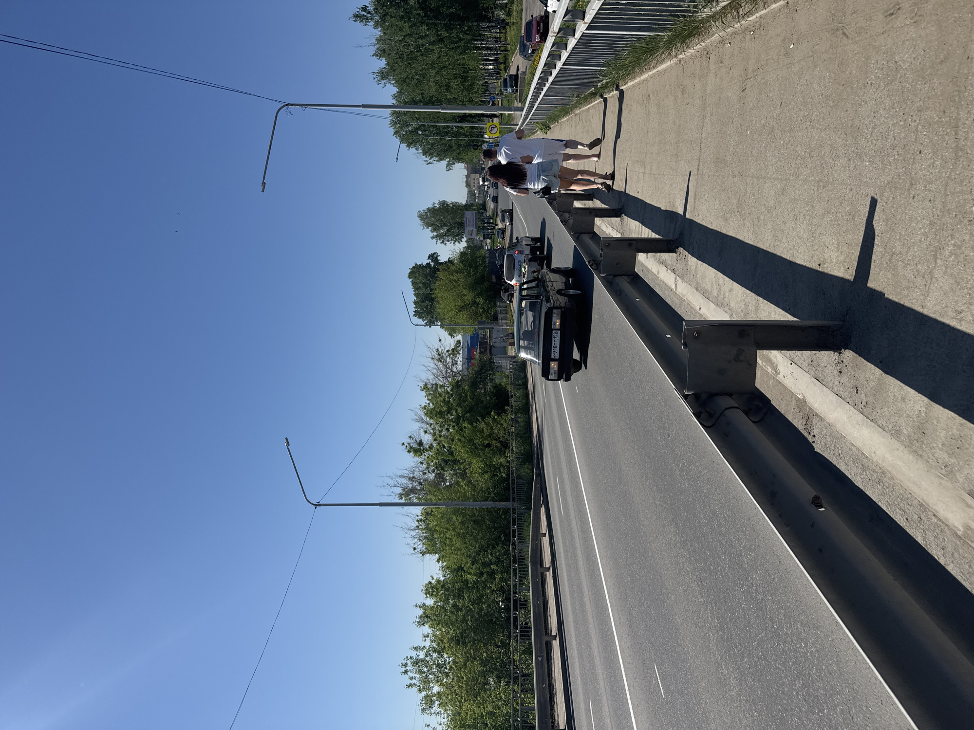 A view from a bridge in the city of Chaikovsky, Russia, shows a two-lane road with a metal guardrail separating it from a pedestrian sidewalk. A black car with license plate number "У 978 ТТ 159" is visible on the road. Two people, one wearing a white top and the other a white dress, are walking on the sidewalk. Streetlights are present along the road. Green trees line the road below the bridge. The sky is clear and blue.