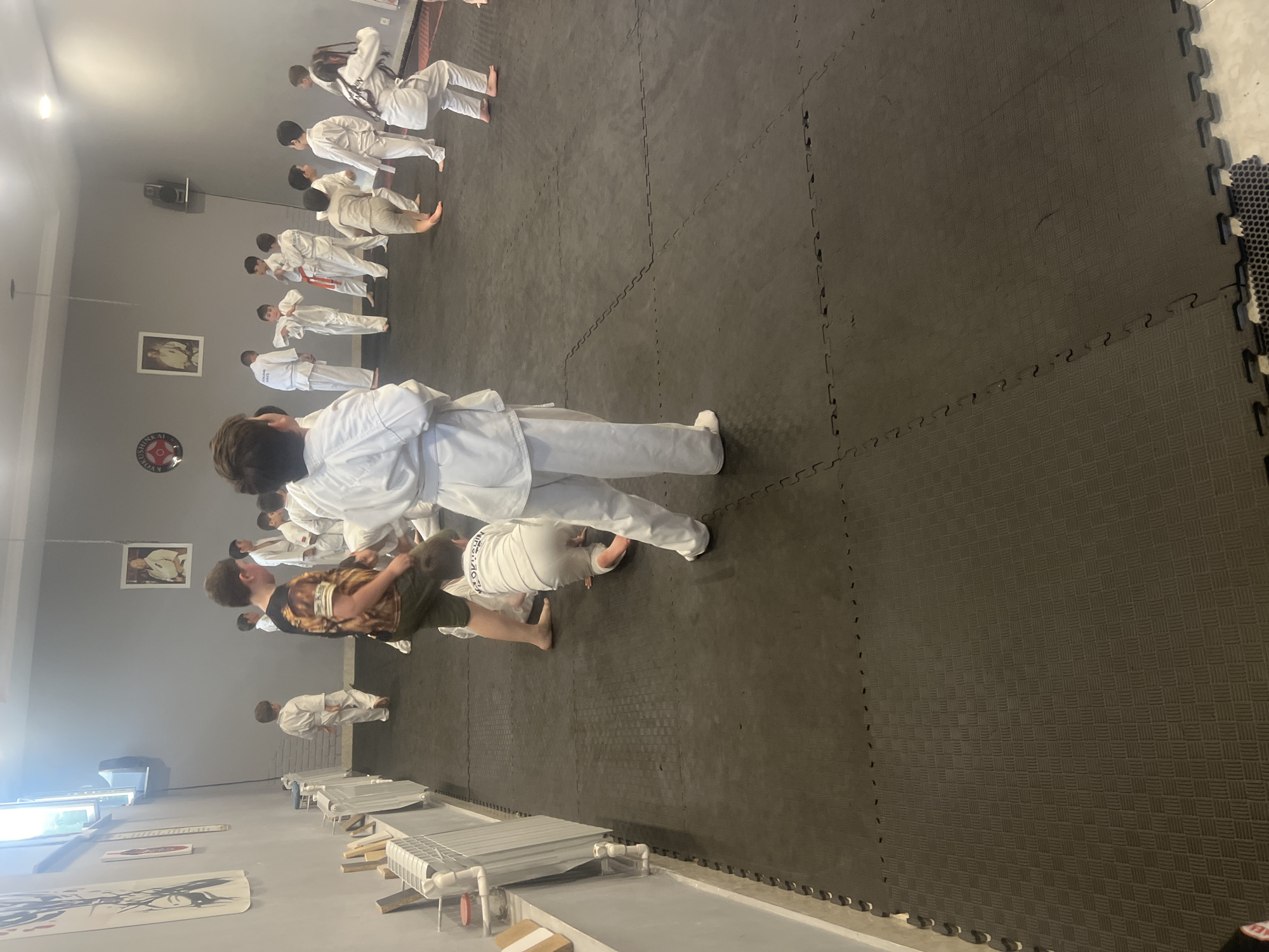 The image depicts a group of people, mostly young individuals dressed in white karate gis, participating in a martial arts class. The scene is an indoor dojo with a dark, interlocking foam mat floor. The lighting suggests it could be daytime, as there are windows visible and interior lights are on. The individuals are in various training poses, suggesting an ongoing class or demonstration.
In the foreground, a person wearing a white gi and a dark patterned t-shirt is bending over. Behind them, more individuals in white gis are lined up, some performing movements. In the background, a larger group of students, also in white gis, are arranged in rows, some facing away from the camera. The dojo appears to be a dedicated training space, with some decorative elements on the walls, including a framed picture and a circular emblem with text. There are also some white racks or benches along the side of the room.
Notable details include the various stances and movements of the karate practitioners, indicating a dynamic activity. The overall atmosphere seems focused and disciplined, typical of martial arts training. The walls are a neutral gray, and the floor provides a cushioned surface for the activity. The presence of multiple students of different ages suggests a community or club setting. Visible text on the circular emblem on the wall appears to include "KYOKUSHIN" and possibly a place name, though it is not fully legible.