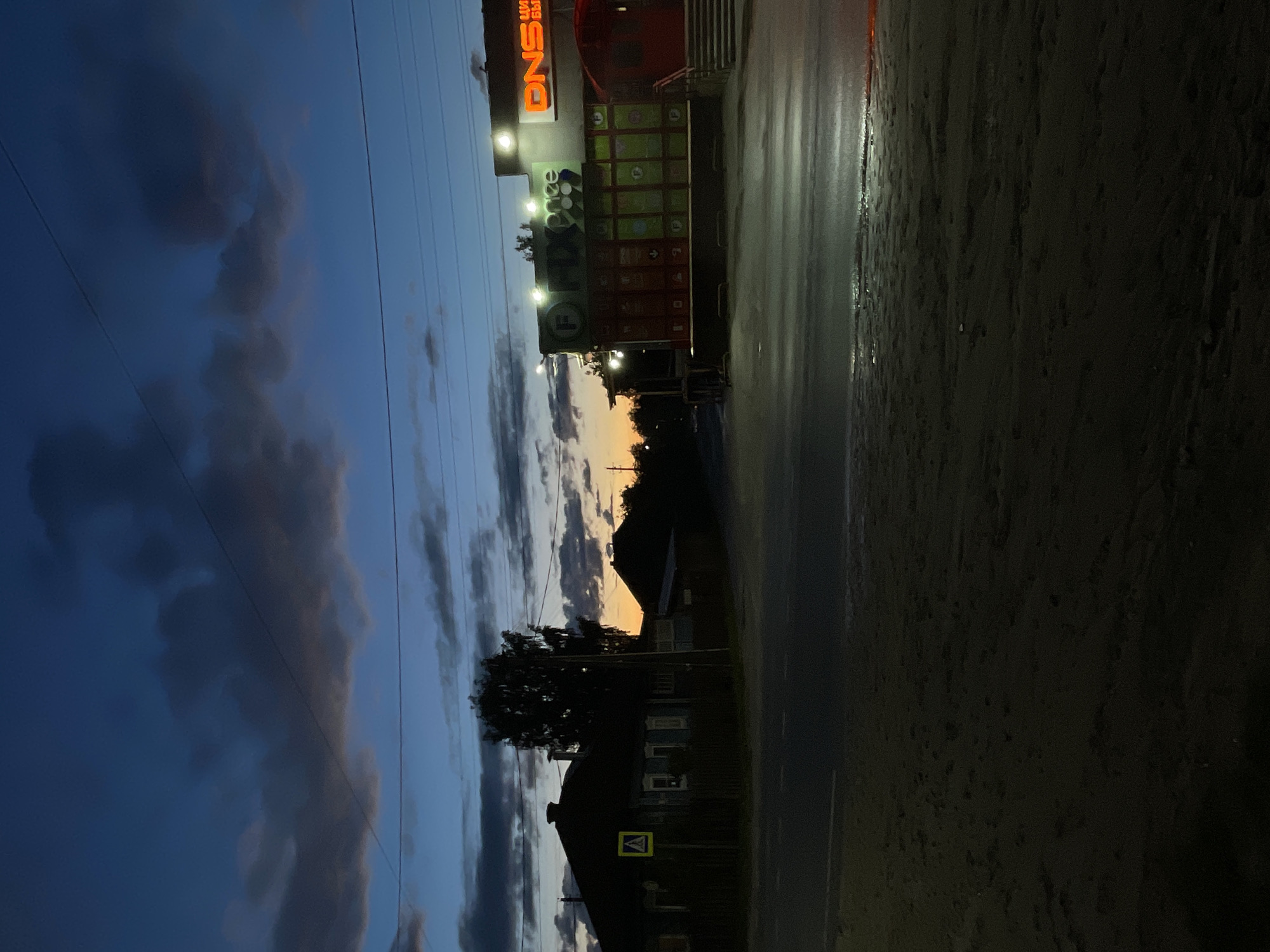 The image depicts a twilight scene, likely dusk or dawn, in Красные Баки, Russia. The sky, dominating the upper left and central portions, transitions from deep blues and purples to a warm orange and yellow glow on the horizon to the right, indicating the time of day. Scattered, dark clouds are visible across the sky.
On the left side of the frame, a large commercial building is brightly illuminated. A prominent orange sign on its facade displays the text "DNS". Above and to the right of "DNS," partial Russian text is visible, and below it, on green-colored panels, letters such as "PD" or "FD" can be discerned. The paved ground directly in front of the building appears wet and reflective, suggesting recent rainfall.
In the middle ground and extending towards the right, the silhouettes of trees and several dark outlines of houses or smaller buildings are visible against the lighter horizon. Near one of these structures, a yellow square sign with a black border and a central black triangle containing an inverted 'V' shape (a standard pedestrian crossing symbol) is visible. Utility lines stretch across the sky in several places.
The scene conveys a quiet, semi-urban or rural atmosphere at the transition between day and night, characterized by a blend of artificial lighting and natural twilight colors.