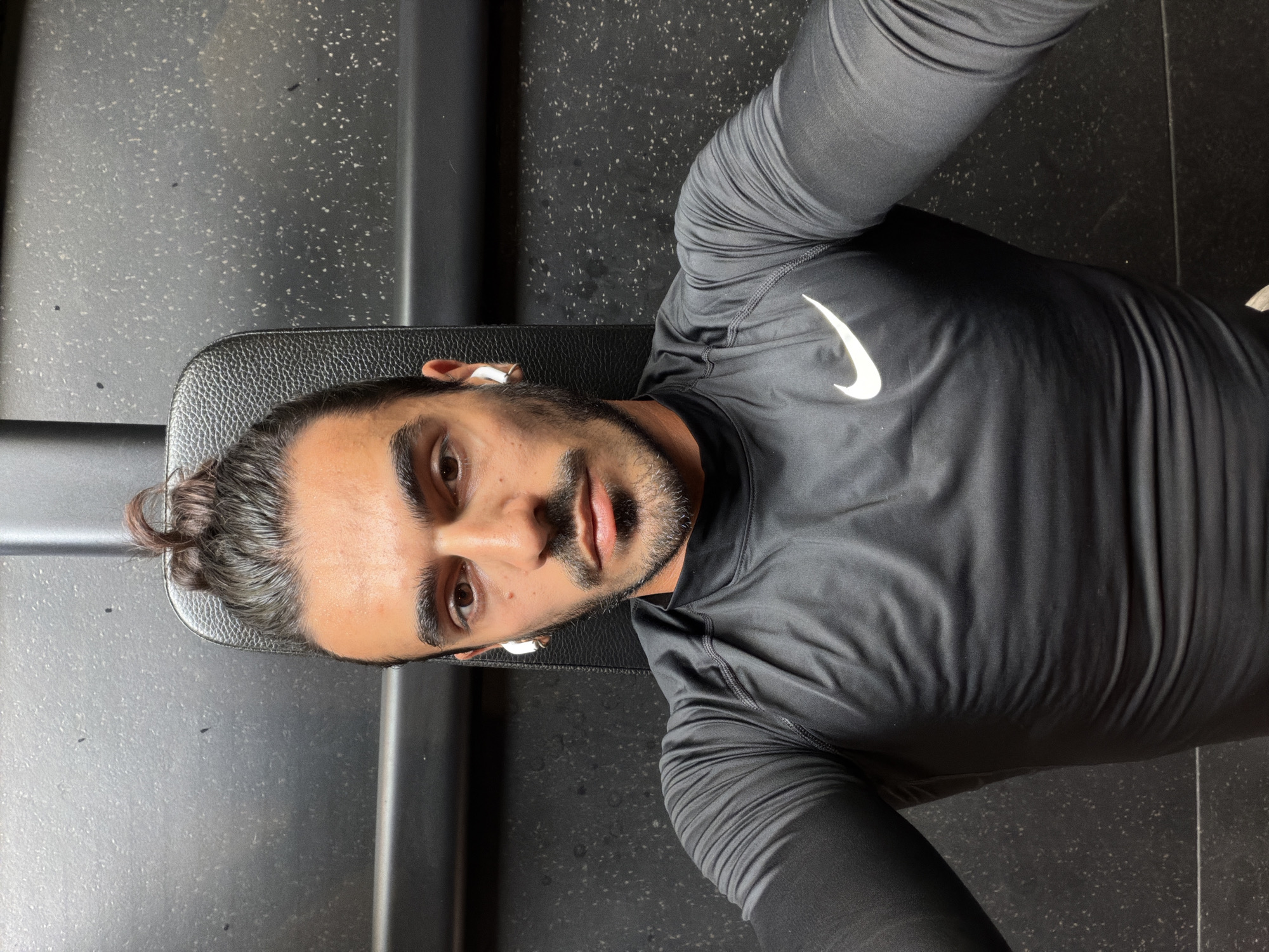 A man lies on a black bench in what appears to be a gym. He is wearing a black long-sleeved athletic shirt with a white Nike swoosh logo on the chest. He has dark hair pulled back in a bun and a short beard. Wireless earbuds are visible in both ears. The floor is black with light speckles. The location is most likely North Kuta, Indonesia.
