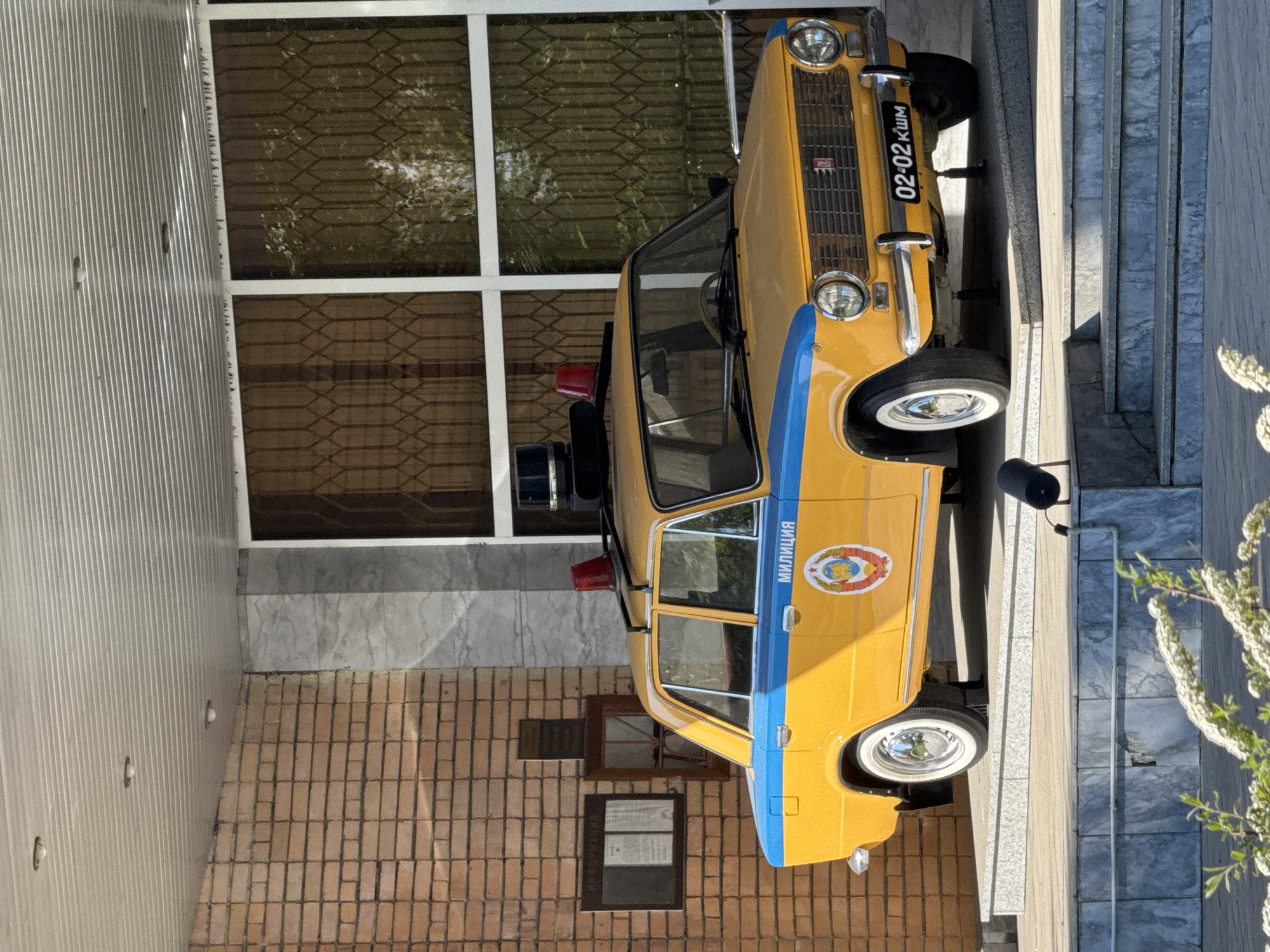 A yellow and blue police car with the word "милиция" on the side is parked next to a building in an unknown location. The car's license plate number is 02-02 КЦМ. The building has a brick facade with windows and a plaque. There are stairs next to the car and a white plant on the right.