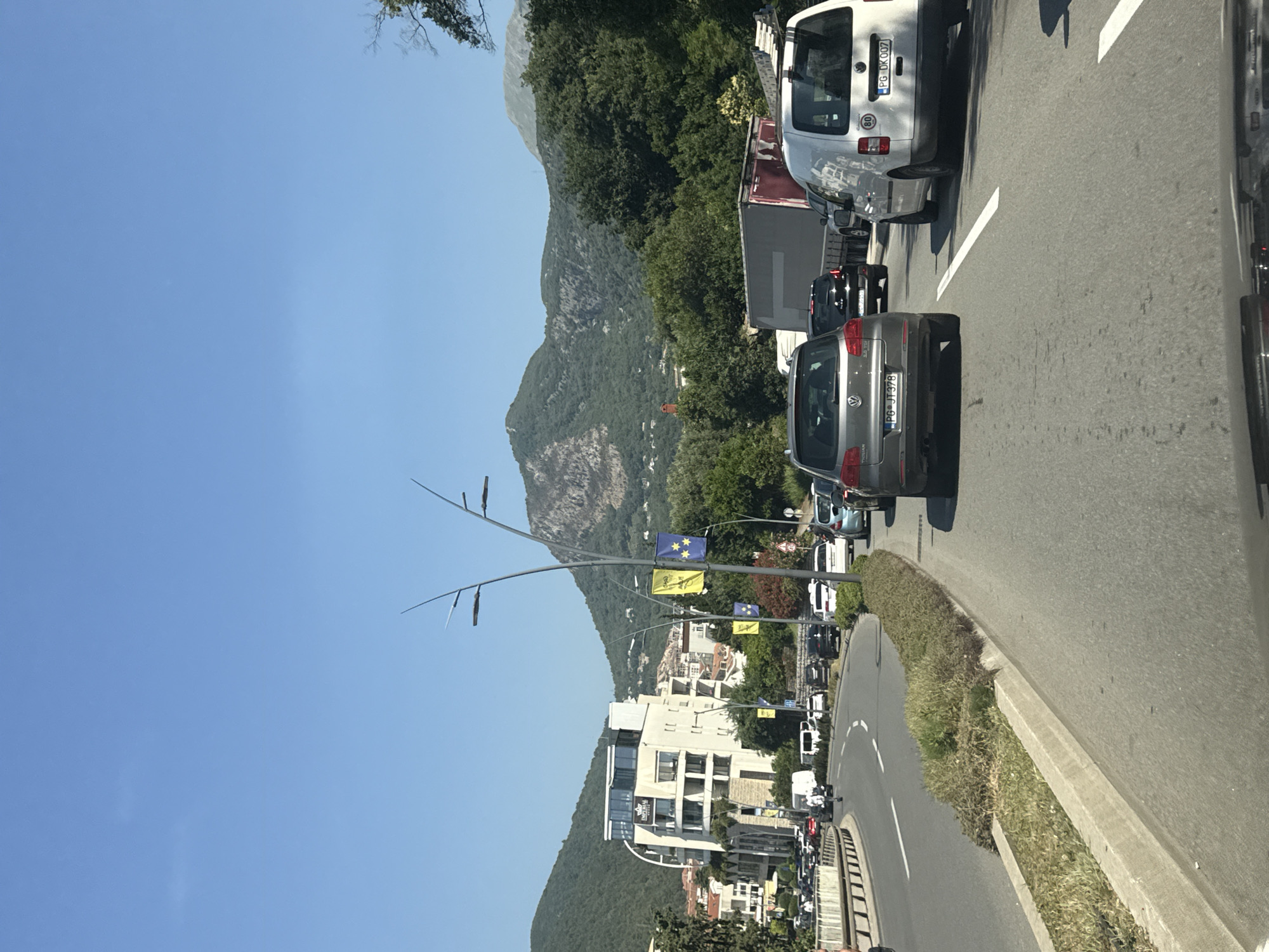 The image captures a traffic jam on a sunny day in Budva, Montenegro. The scene is dominated by a road with several cars, including a silver Volkswagen Tiguan with license plate PG JT 378, a black car, and a white van with license plate PG DK 007. In the background, a large building with "Imperial" written on it is visible, along with other buildings and lush green mountains. Streetlights with decorative flags adorn the roadside. The sky is clear blue.