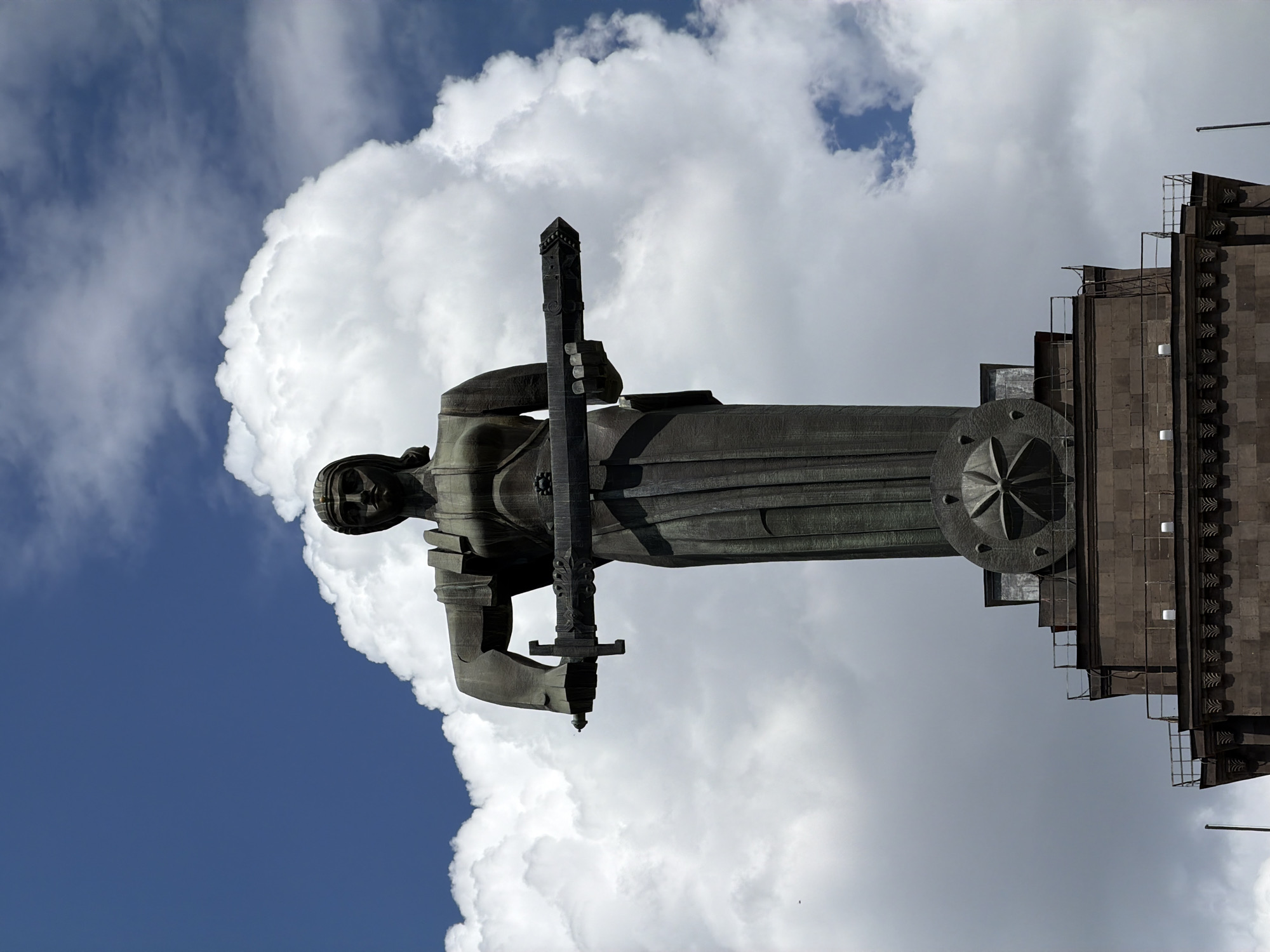 The image shows the Mother Armenia statue in Yerevan, Armenia, against a backdrop of puffy white clouds and blue sky. The statue is made of dark metal and depicts a woman holding a sword. The statue stands on a multi-tiered stone base with a decorative element resembling a rosette. The statue and the base are positioned on an elevated location, offering a view of the sky behind. The orientation of the image is portrait, rotated 90 degrees clockwise.