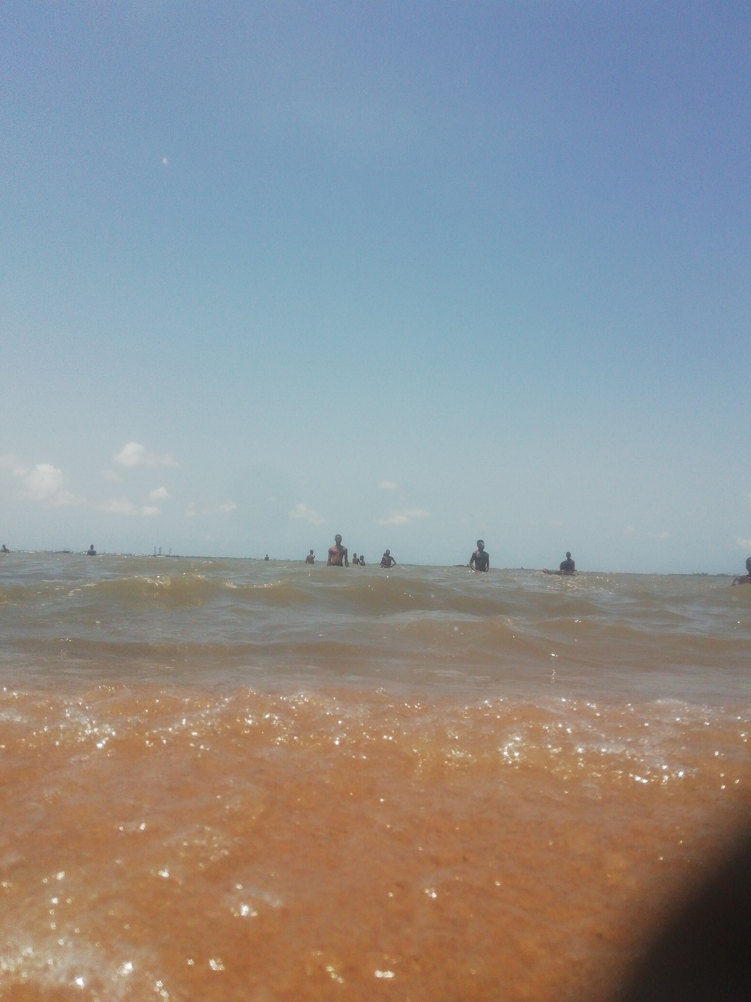 The image shows a beach scene in Ikorodu, Nigeria. The foreground is dominated by the wet, brownish-orange sand of the beach. The water is murky and brown, with small waves rippling across the surface. In the distance, several people are standing or swimming in the water. The sky is blue and clear, with a few scattered clouds. The image is taken from a low angle, giving the viewer a sense of being close to the water.